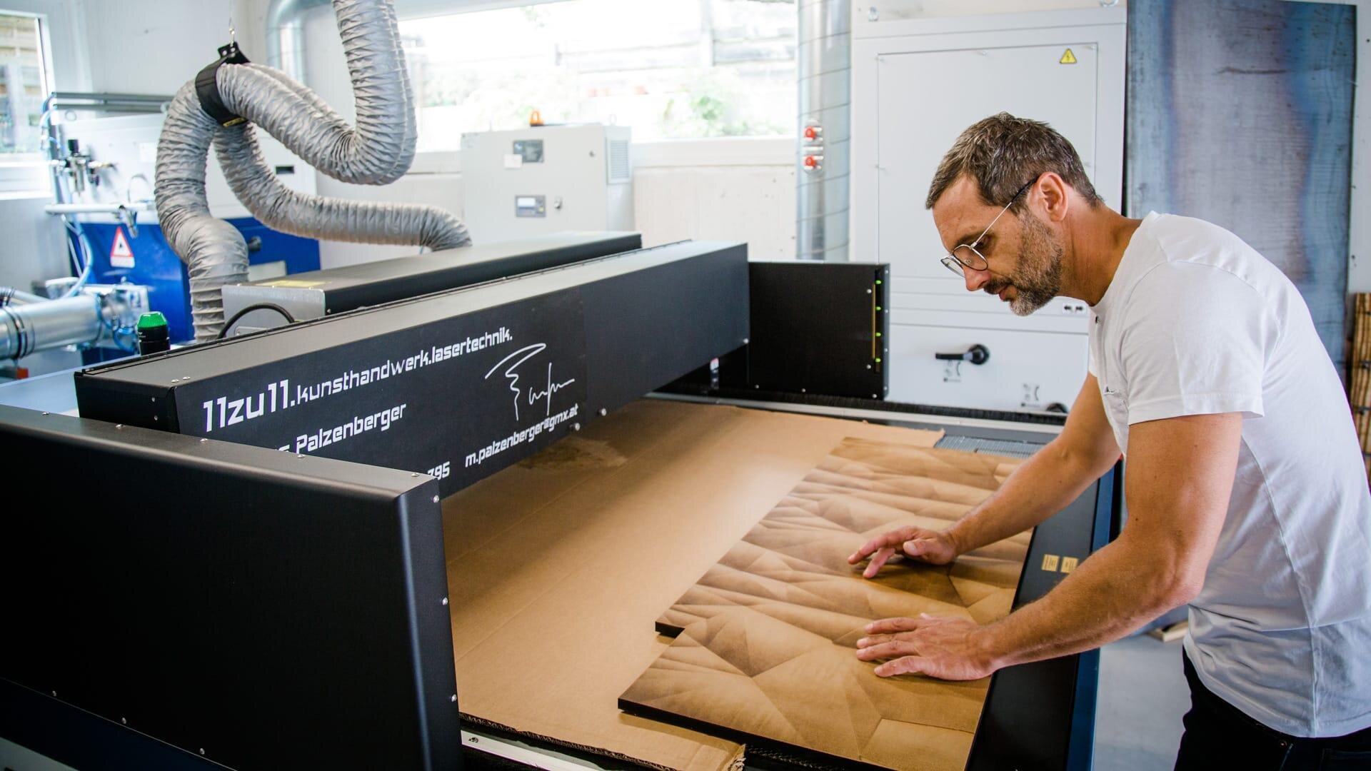 Ein Mann mit Brille und weißem Hemd prüft eine Holzplatte mit einem in 3D geschnitzten Muster. Er arbeitet an einer großen schwarzen CNC-Maschine in einer hellen, modernen Werkstatt.