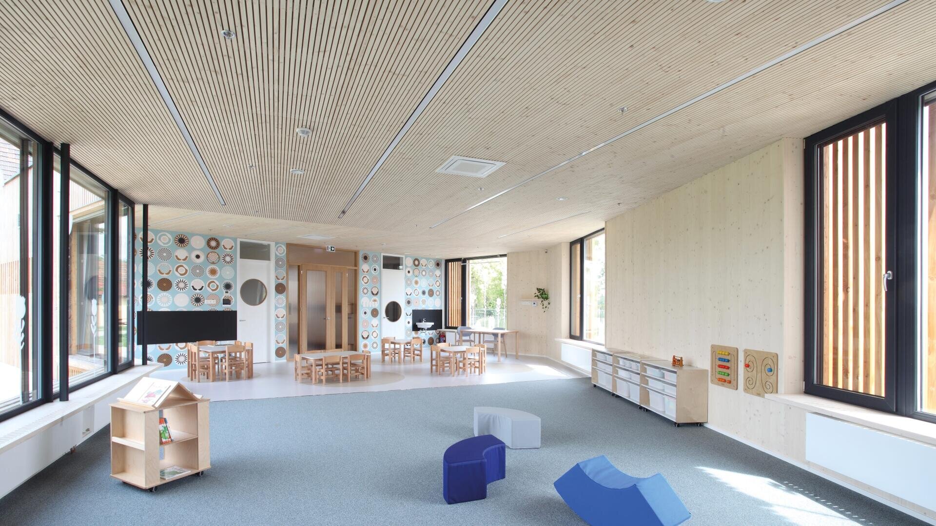Ein helles, geräumiges Klassenzimmer mit großen Fenstern, Holzwänden und -decken, kleinen Tischen und Stühlen, Lernspielzeug, Regalen und bunten, geometrischen Sitzblöcken auf einer Teppichfläche.