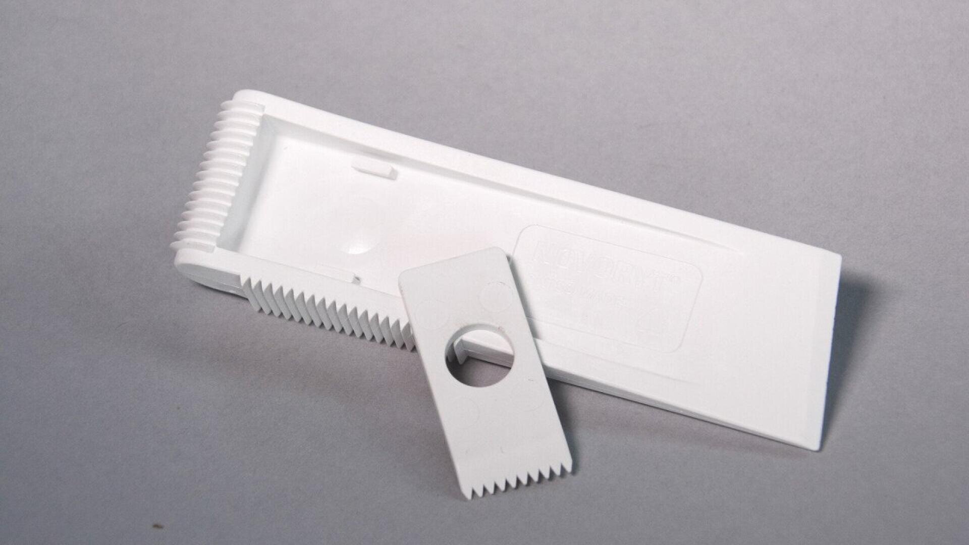 Peigne à poux en plastique blanc avec des dents très rapprochées et une partie amovible comportant un trou, présenté sur un fond gris uni.
