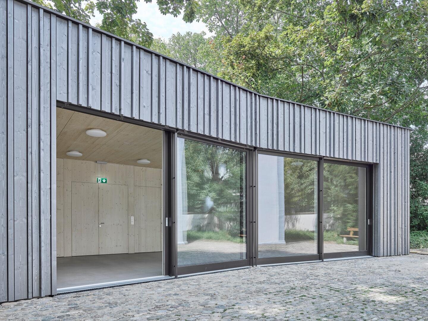 Ein modernes Gebäude mit vertikaler Holzverkleidung, großen, raumhohen Glastüren und einem hellen Holzinterieur. Die Türen öffnen sich zu einem gepflasterten Innenhof und geben den Blick frei auf Grünflächen und Bäume im Freien.