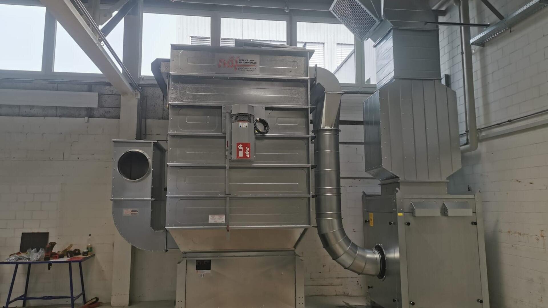 Großes industrielles Luftfiltersystem aus Metall, das in einem Innenraum vor einer weißen Backsteinwand mit Fenstern darüber installiert ist. Das System besteht aus Kanälen und einem Schaltkasten mit einem Feuerlöscher an der Vorderseite.