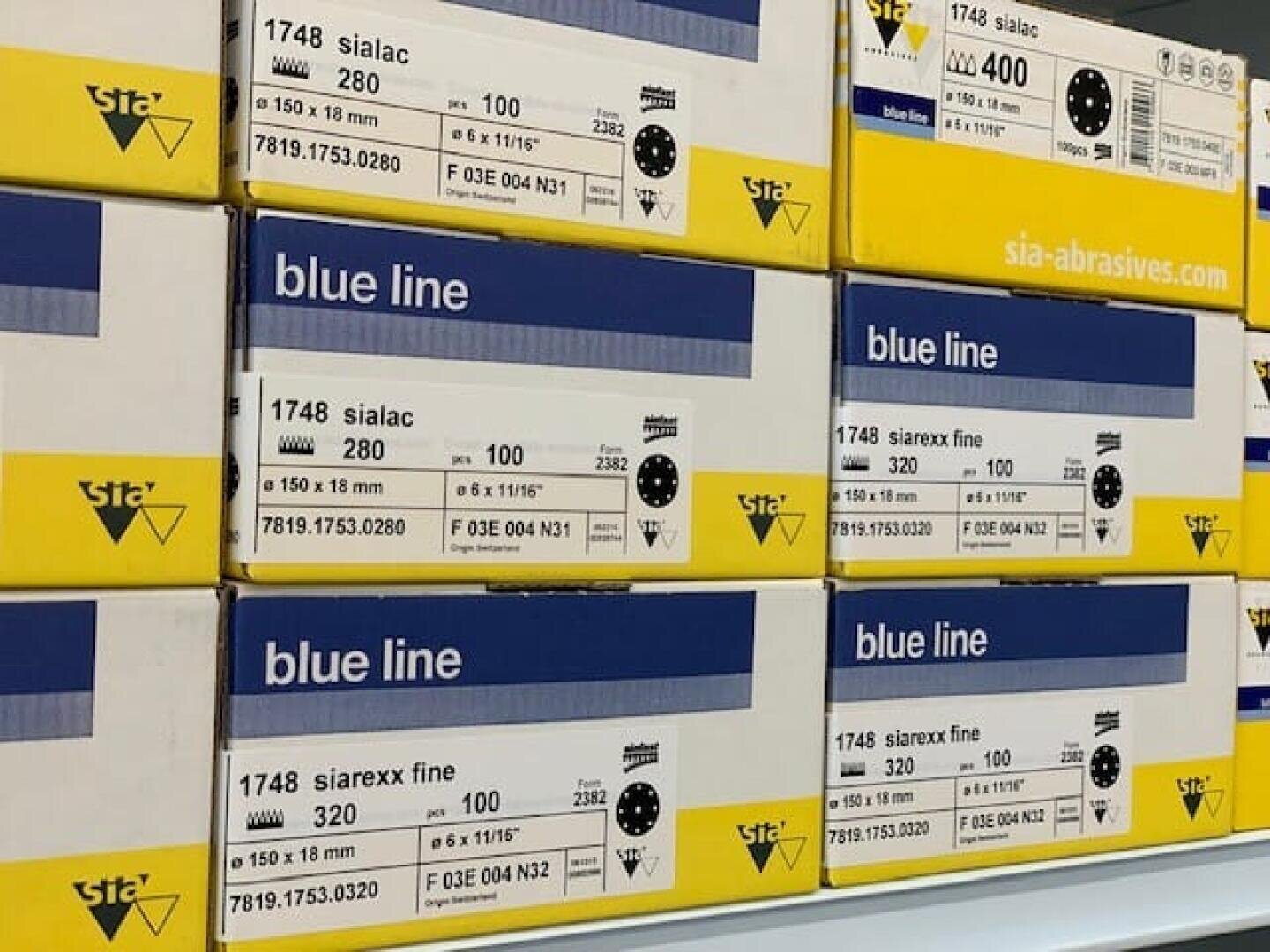 Gestapelte Schachteln von Sia-Schleifmitteln, mit blauer Linie beschriftet, mit Angaben wie Korngröße (280 und 320), Abmessungen (150 x 118 mm) und Produktcodes auf der Verpackung.