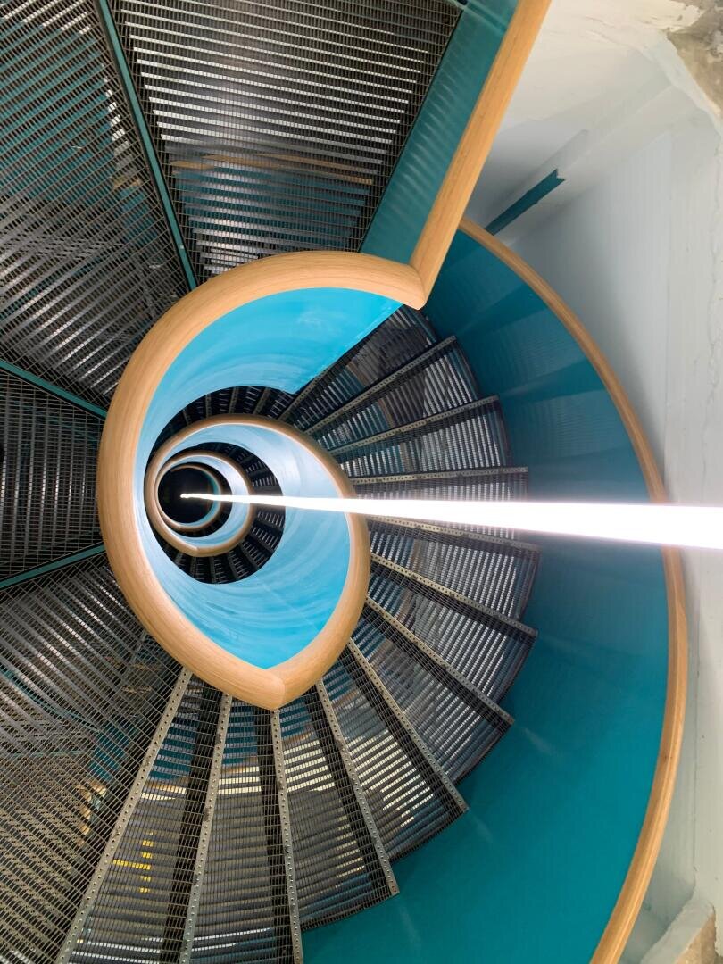 Spindeltreppe mit türkisfarbenen Wänden, Metallstufen und einem hölzernen Handlauf, von oben gesehen, die ein geometrisches, hypnotisches Muster mit einem zentralen weißen vertikalen Licht erzeugt.