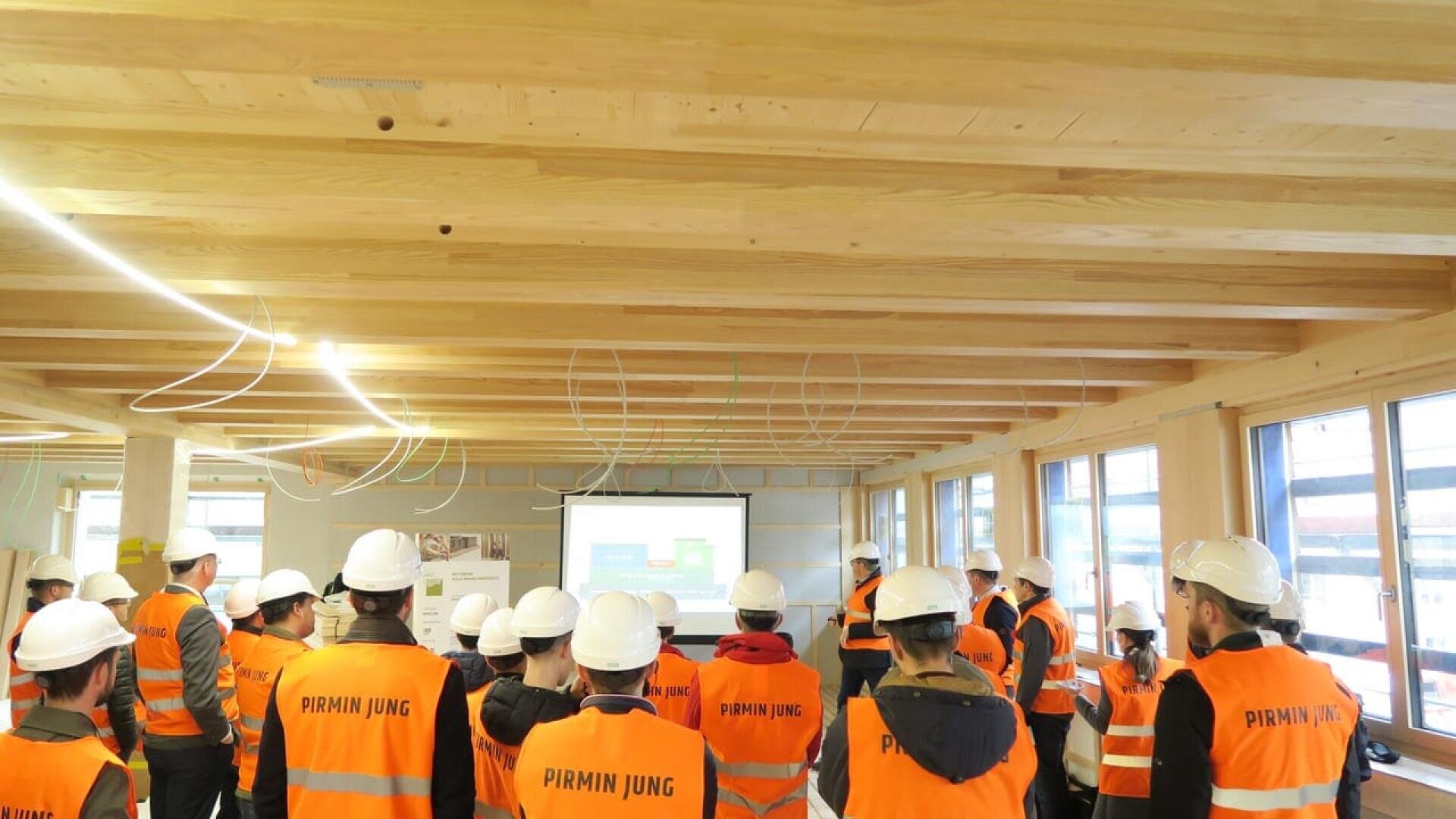 Un groupe de personnes portant des casques blancs et des gilets de sécurité orange étiquetés PIRMIN JUNG assistent à une présentation dans un bâtiment en bois en construction, face à un écran de projecteur situé à l'avant de la pièce.