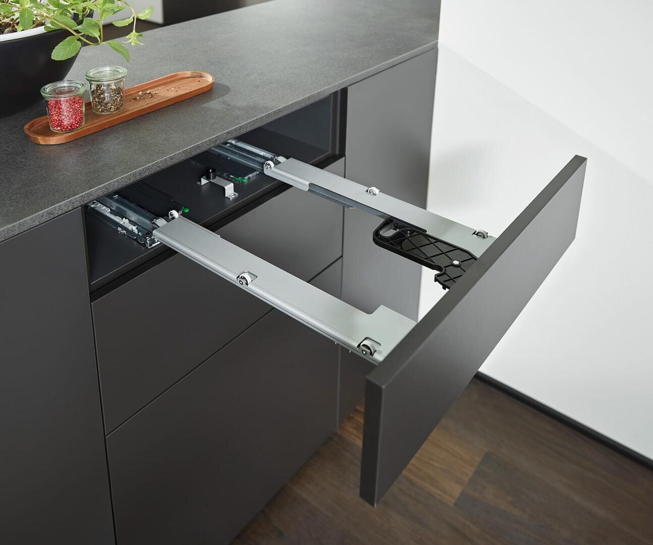 Ein moderner schwarzer Küchenschrank mit einem ausziehbaren, klappbaren Regal, das teilweise ausgezogen ist und Metallschiebevorrichtungen aufweist; auf der grauen Arbeitsplatte darüber stehen Gewürzdosen und eine Pflanze.