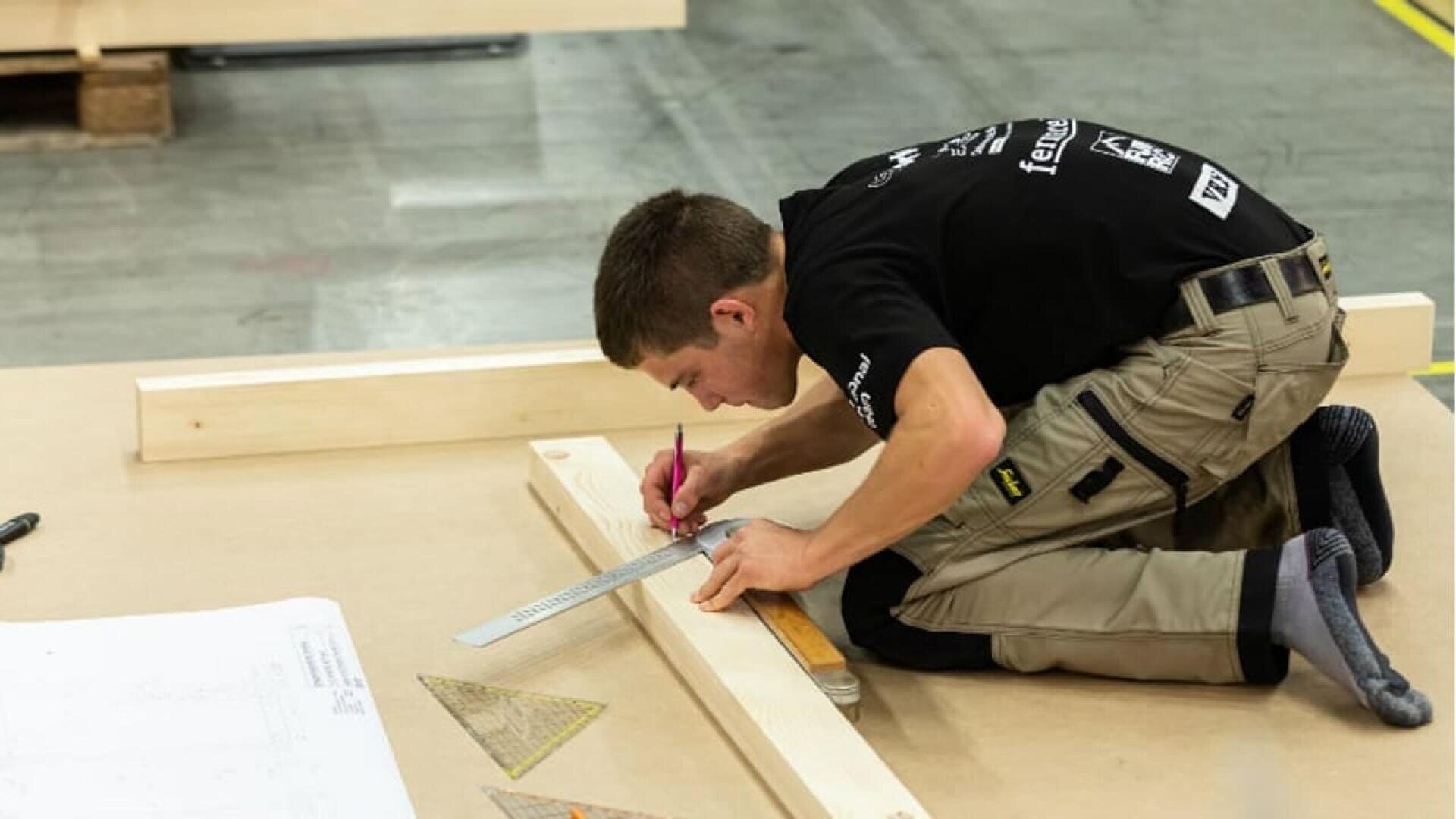 Une personne est agenouillée sur le sol et utilise une règle et un crayon pour marquer des mesures sur une planche de bois. D'autres planches de bois, un triangle de mesure et des plans en papier se trouvent à proximité. Le décor semble être celui d'un atelier ou d'une zone de construction.