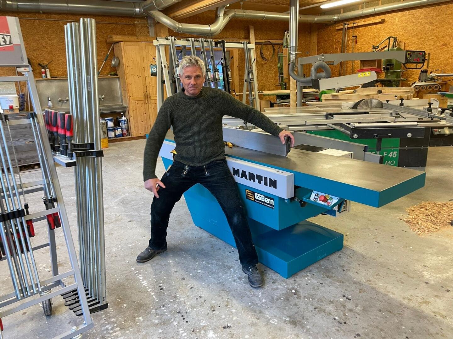 Ein Mann in einer Werkstatt posiert mit einem Bein auf jeder Seite einer großen grünen MARTIN Holzbearbeitungsmaschine. Im Hintergrund sind verschiedene Werkzeuge und Holzbearbeitungsgeräte auf einem Holzboden zu sehen.