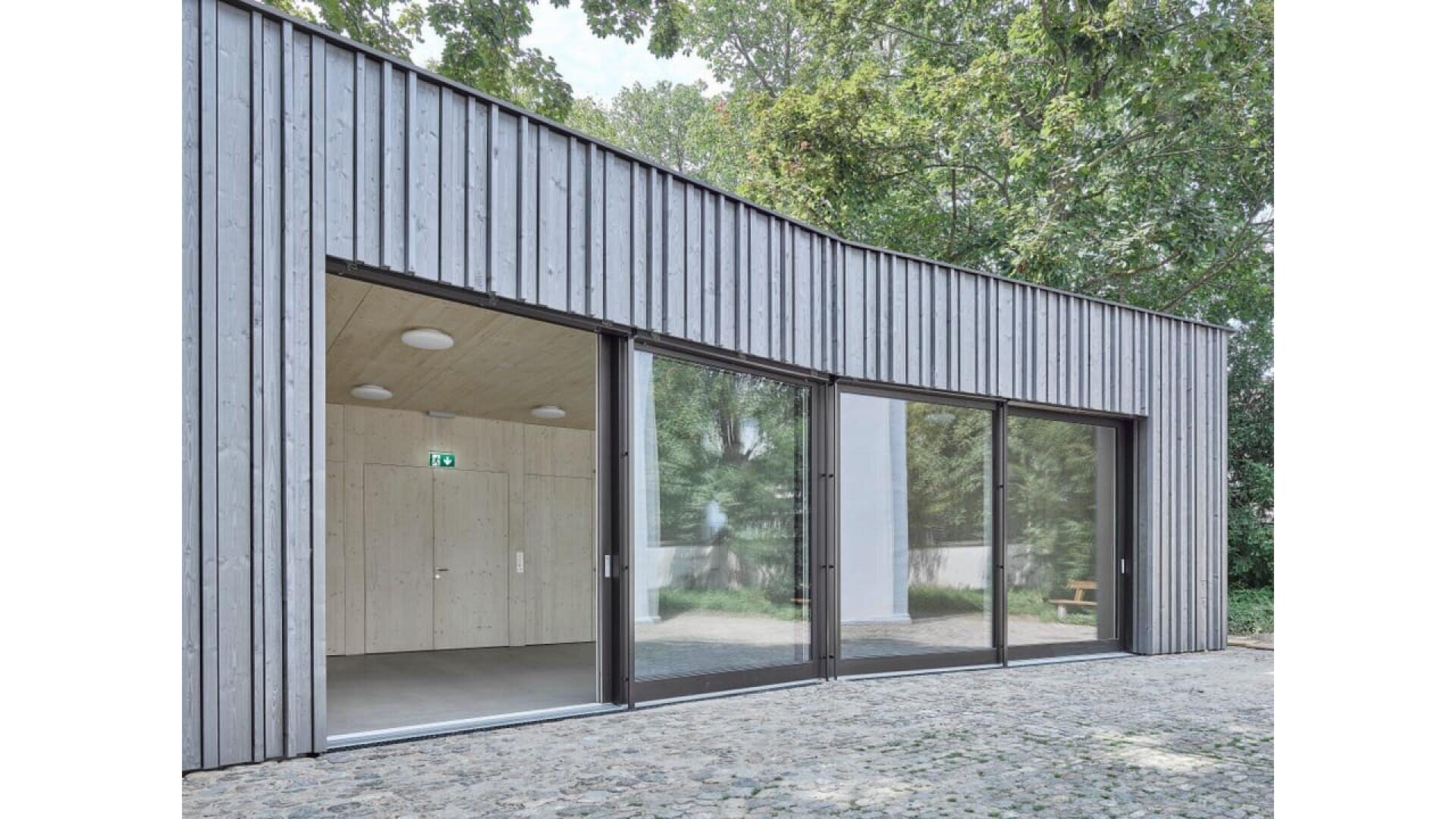 Bâtiment moderne avec bardage en bois vertical, grandes portes coulissantes en verre du sol au plafond et intérieur en bois clair, entouré d'arbres et d'un patio en pavés.