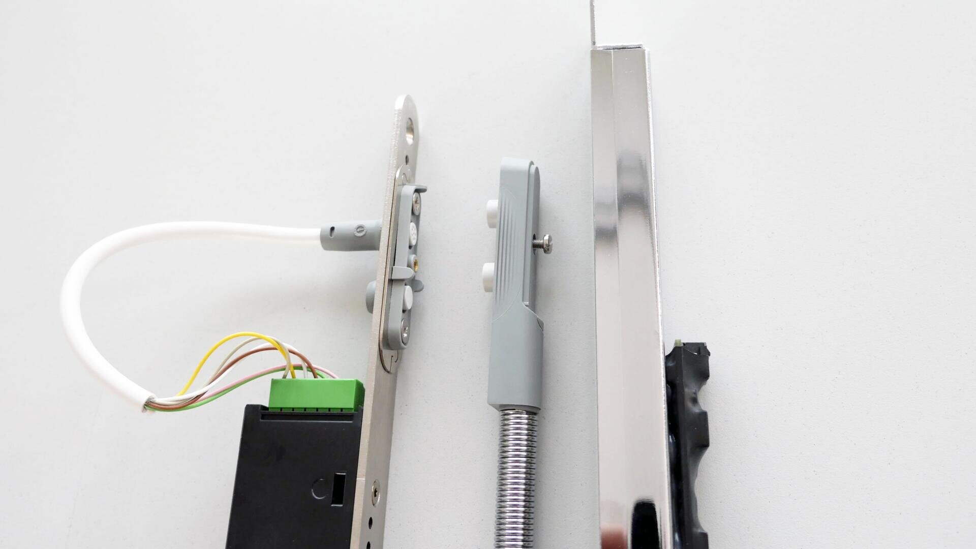 Drei metallische Tür- oder Fensterbeschlagteile, darunter ein Schlossmechanismus mit angebrachten Drähten, ein federbelasteter Arm und ein glänzender rechteckiger Stab, sind auf einer weißen Fläche angeordnet.