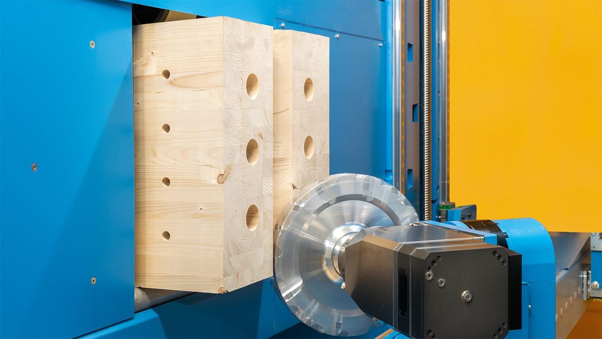 Nahaufnahme einer CNC-Maschine, die Löcher in zwei gestapelte Holzblöcke bohrt, wobei im Hintergrund blaue und gelbe Maschinenteile zu sehen sind.
