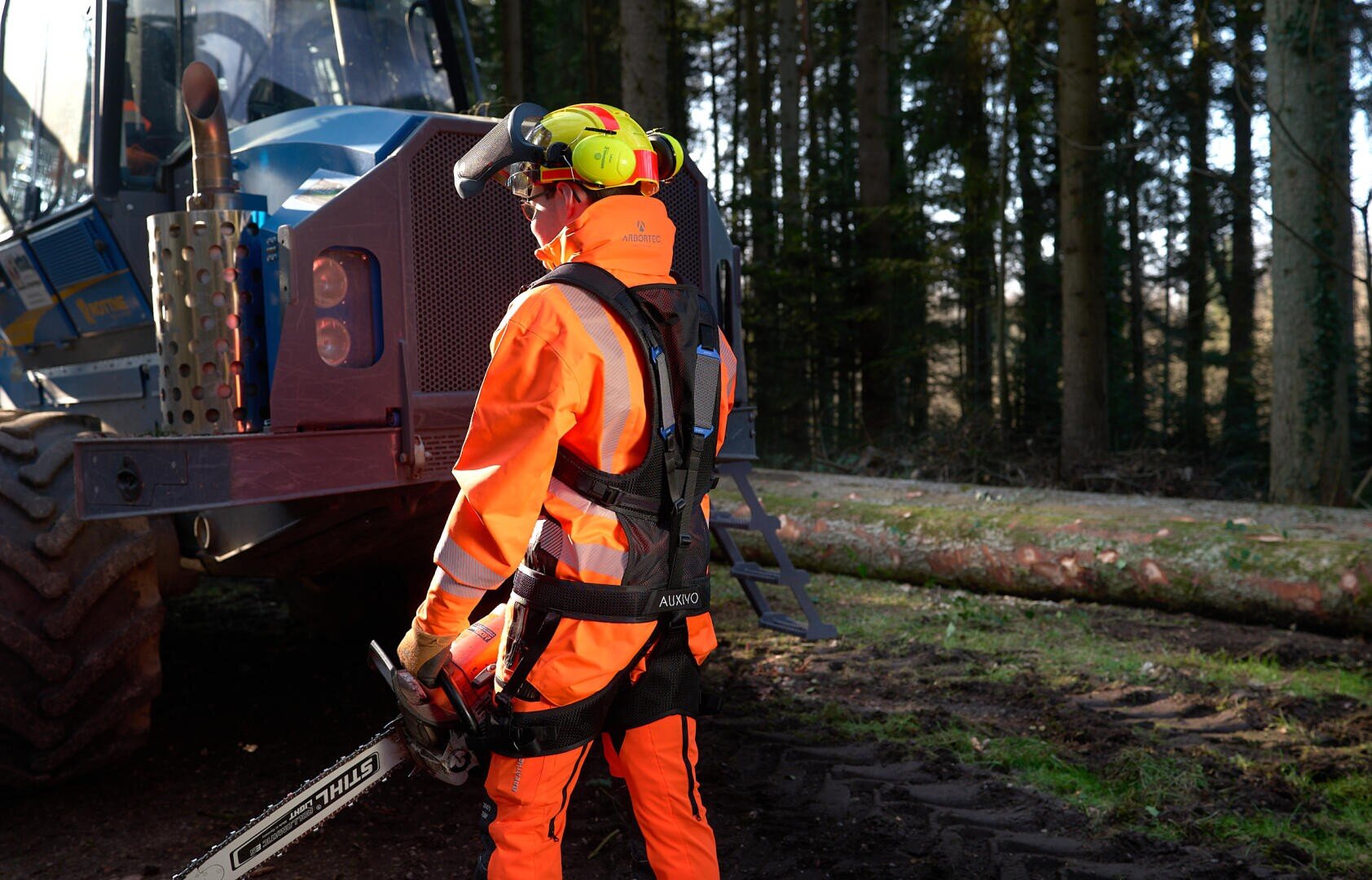 Eine Person in leuchtend orangefarbener Schutzausrüstung und mit Helm steht vor Forstmaschinen und hält eine Kettensäge in einem Waldstück, auf dessen Boden umgestürzte Baumstämme sichtbar sind.