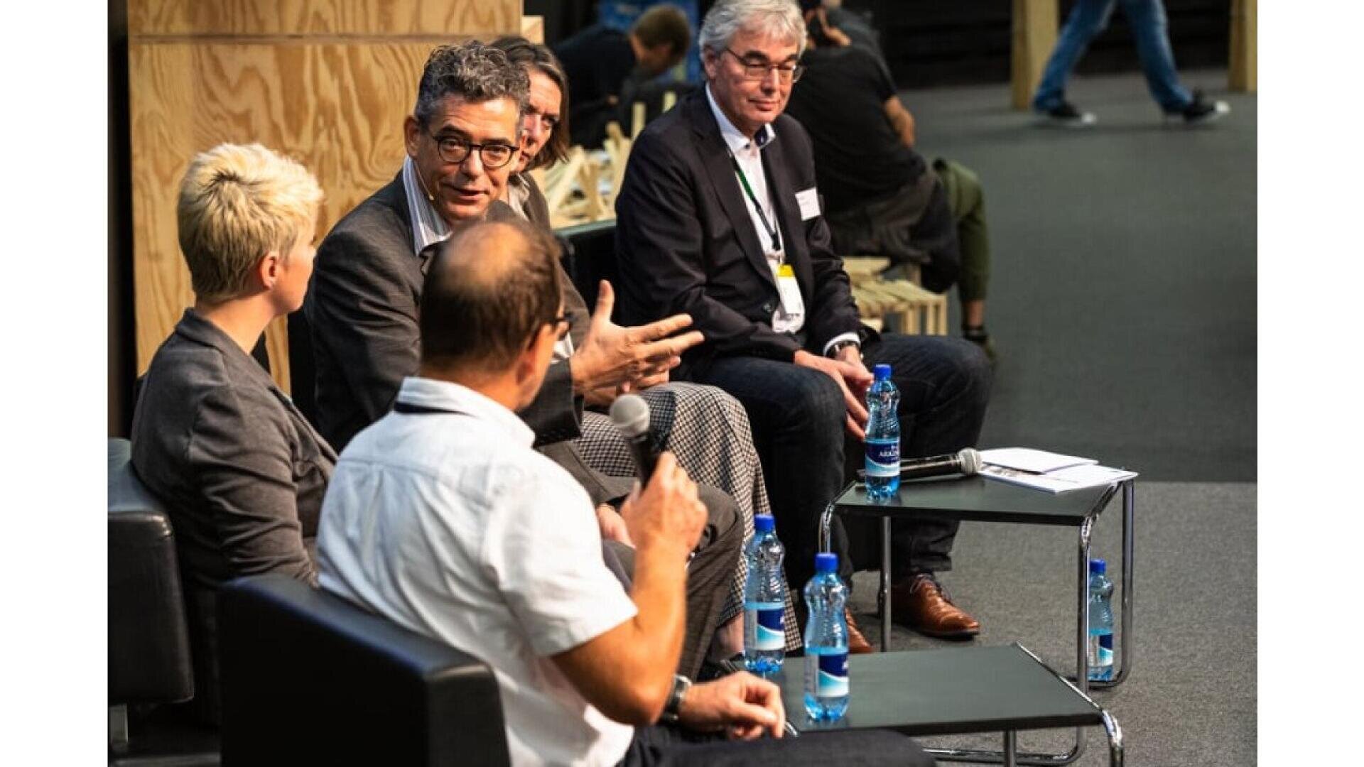 Un groupe de cinq personnes en tenue de travail est assis dans un groupe de discussion, tenant des microphones et discutant. Des bouteilles d'eau et des documents sont posés sur des tables devant eux. Le cadre semble être une salle de conférence ou de séminaire.