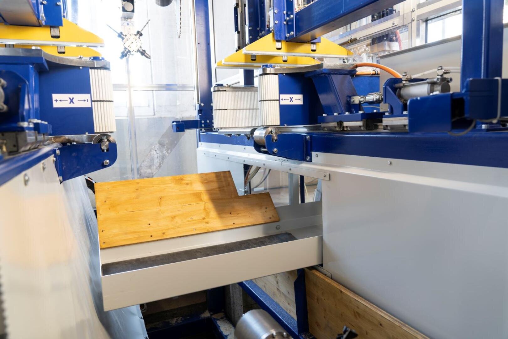 Nahaufnahme einer automatisierten Industriemaschine, die ein Holzbrett bearbeitet. Um den Arbeitsbereich herum sind blaue und gelbe Metallteile, Rollen und Schutzvorrichtungen zu sehen.
