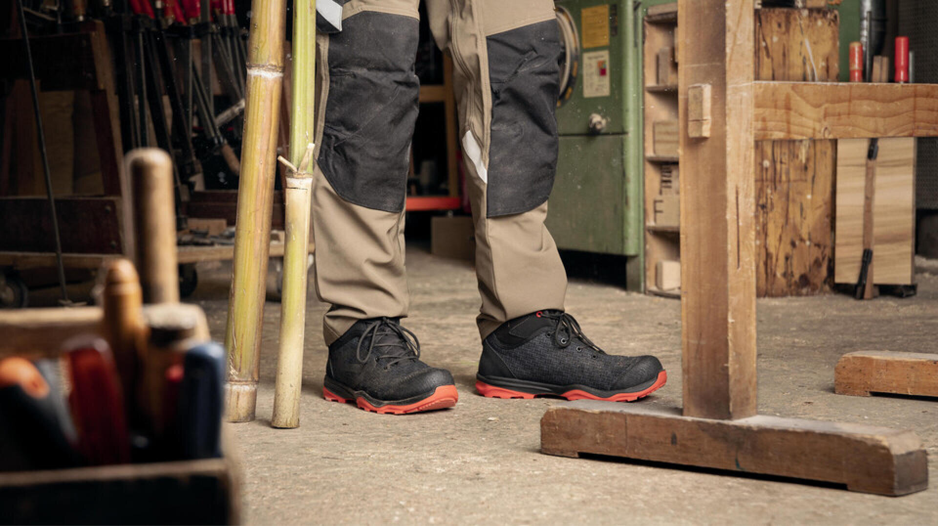 Une personne portant un pantalon de travail beige et noir et des chaussures de sécurité noires à semelles rouges se tient dans un atelier entouré d'outils et d'équipements en bois.