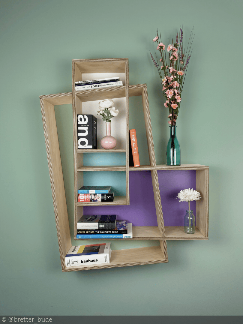 Ein modernes Wandregal aus Holz mit asymmetrischem Design enthält Bücher, eine rosa Vase mit weißen Blumen, eine grüne Vase mit rosa Blumen und eine kleine Glasvase mit einer weißen Blume vor einer grünen Wand.