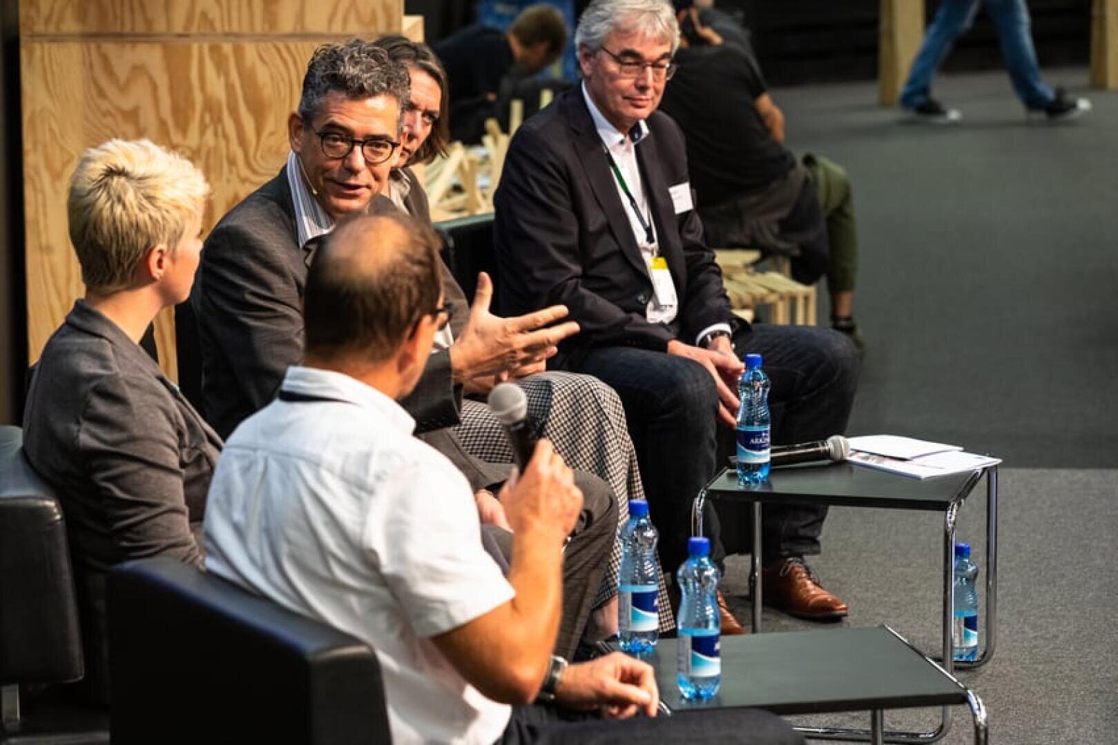 Fünf Personen sitzen bei einer Podiumsdiskussion auf der Bühne und führen ein Gespräch. Sie sitzen auf Stühlen mit Mikrofonen und Wasserflaschen auf kleinen Tischen und blicken auf das Publikum. Der Schauplatz scheint eine Konferenz oder ein Seminar zu sein.