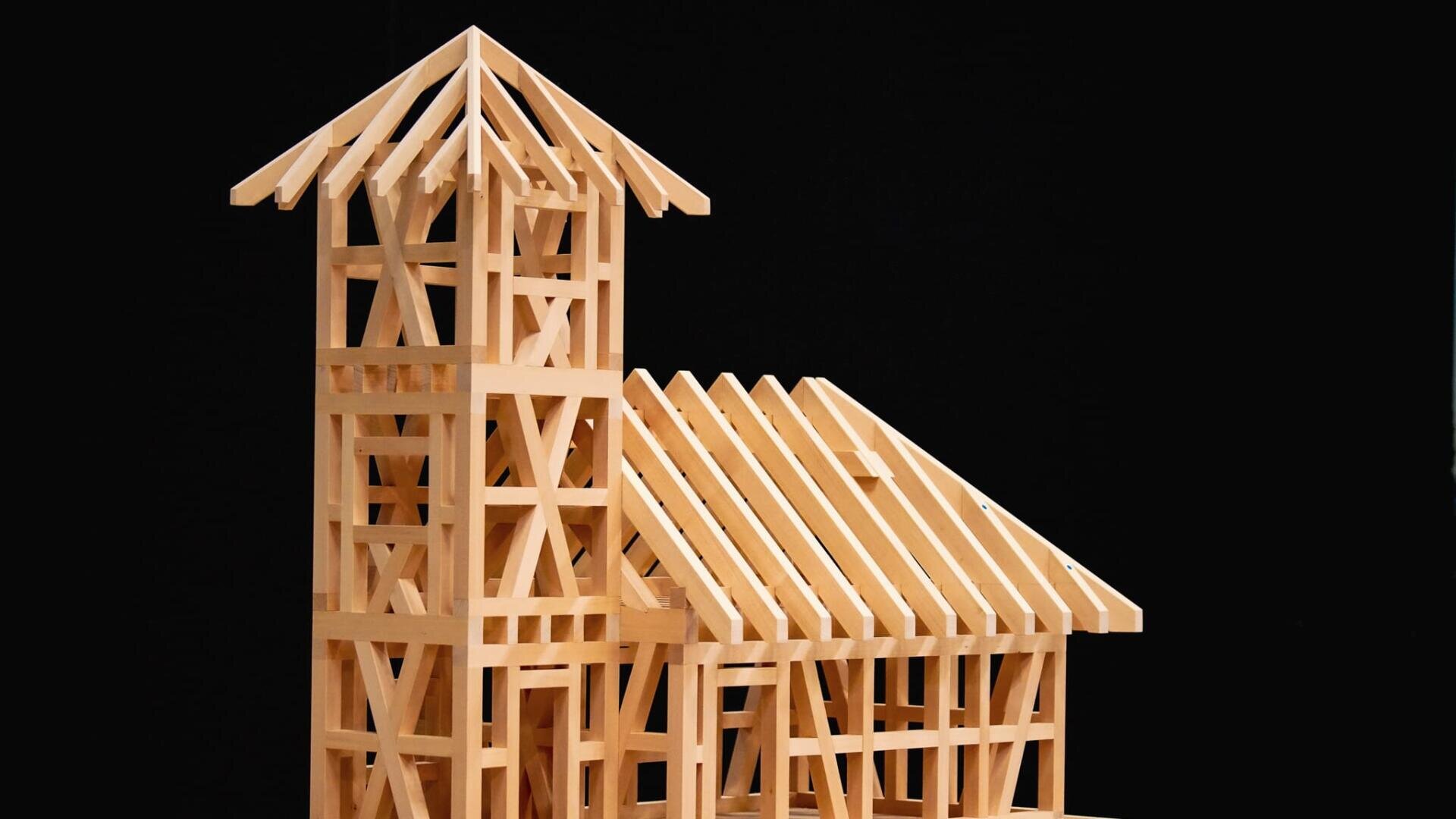 Ein Modell eines Holzhauses mit einem hohen, turmähnlichen Teil und einer schrägen Dachkonstruktion, dargestellt vor einem schwarzen Hintergrund.