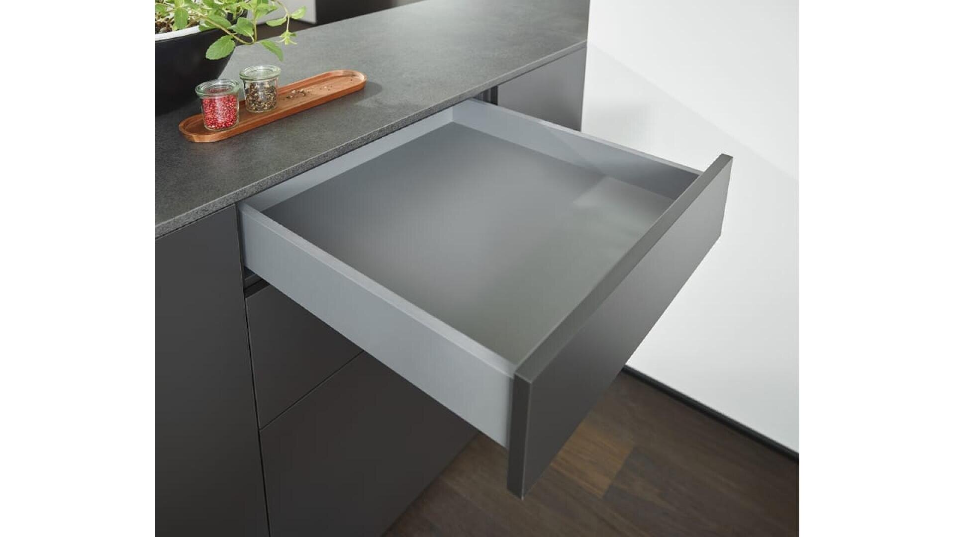 Un tiroir de cuisine moderne, fini en gris mat, est ouvert sous un comptoir noir. Un plateau en bois contenant deux petits pots à épices est posé sur le comptoir près d'une plante verte.