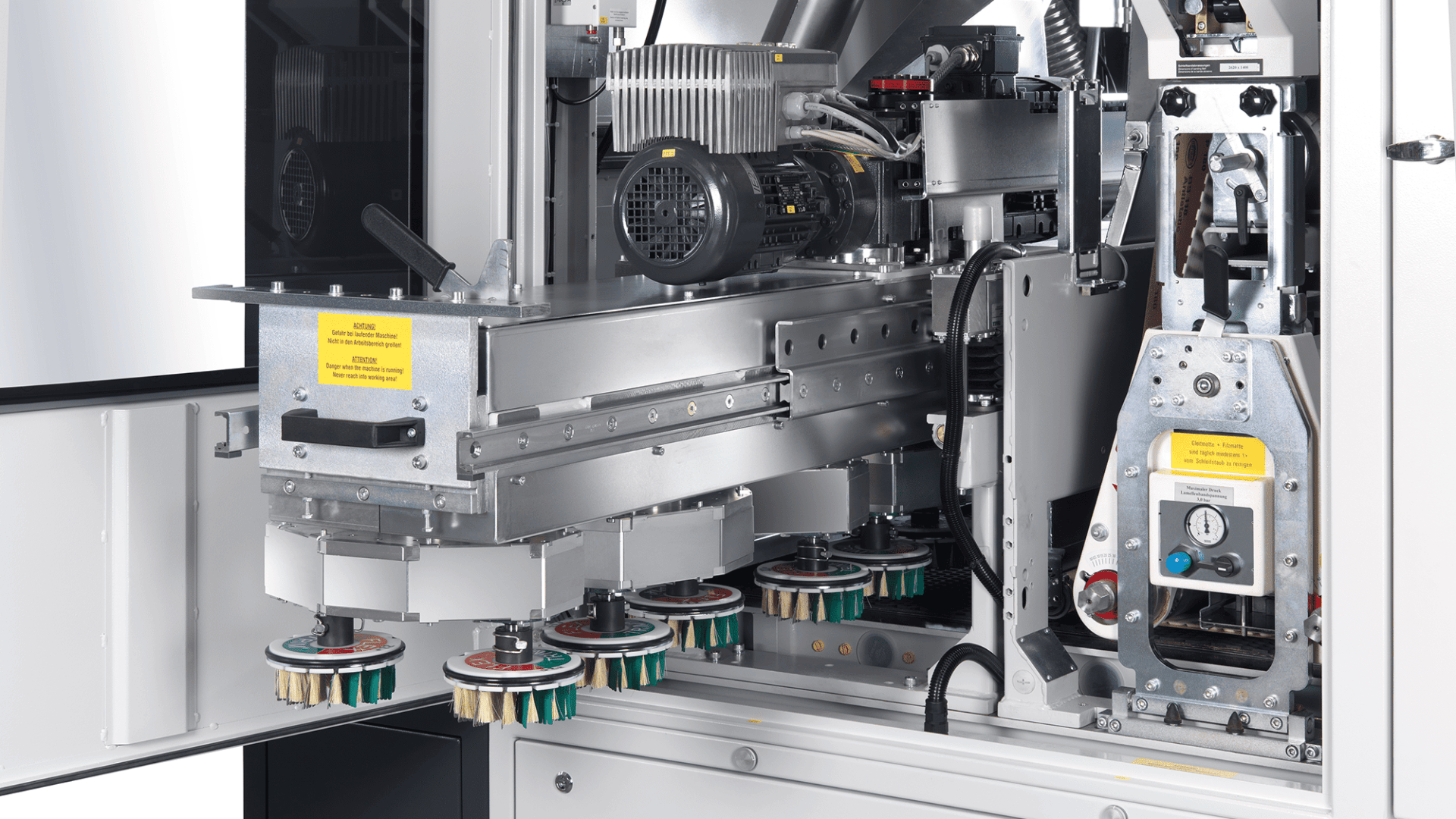Nahaufnahme einer Industriemaschine mit Metallteilen, Motoren, Kabeln und rotierenden Bürstenköpfen, die mit Etiketten und Sicherheitshinweisen versehen sind. Die Ausrüstung scheint für automatische Reinigungs- oder Endbearbeitungsprozesse konzipiert zu sein.