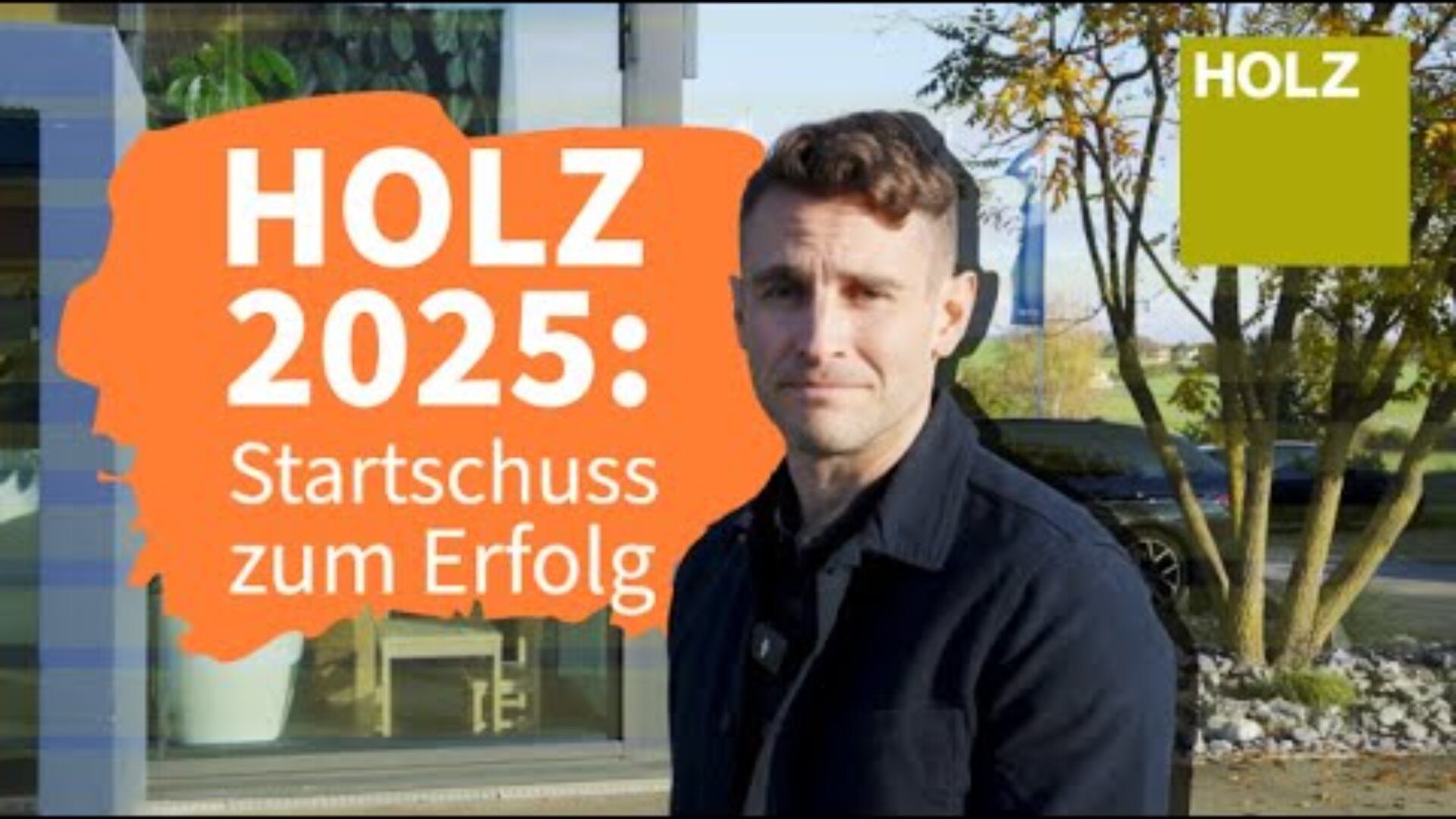 Un homme se tient à l'extérieur, près d'un bâtiment moderne, à côté d'un panneau orange portant l'inscription HOLZ 2025 : Startschuss zum Erfolg. Un arbre et une zone herbeuse sont visibles à l'arrière-plan ; le mot HOLZ apparaît dans un cadre vert dans le coin supérieur droit.