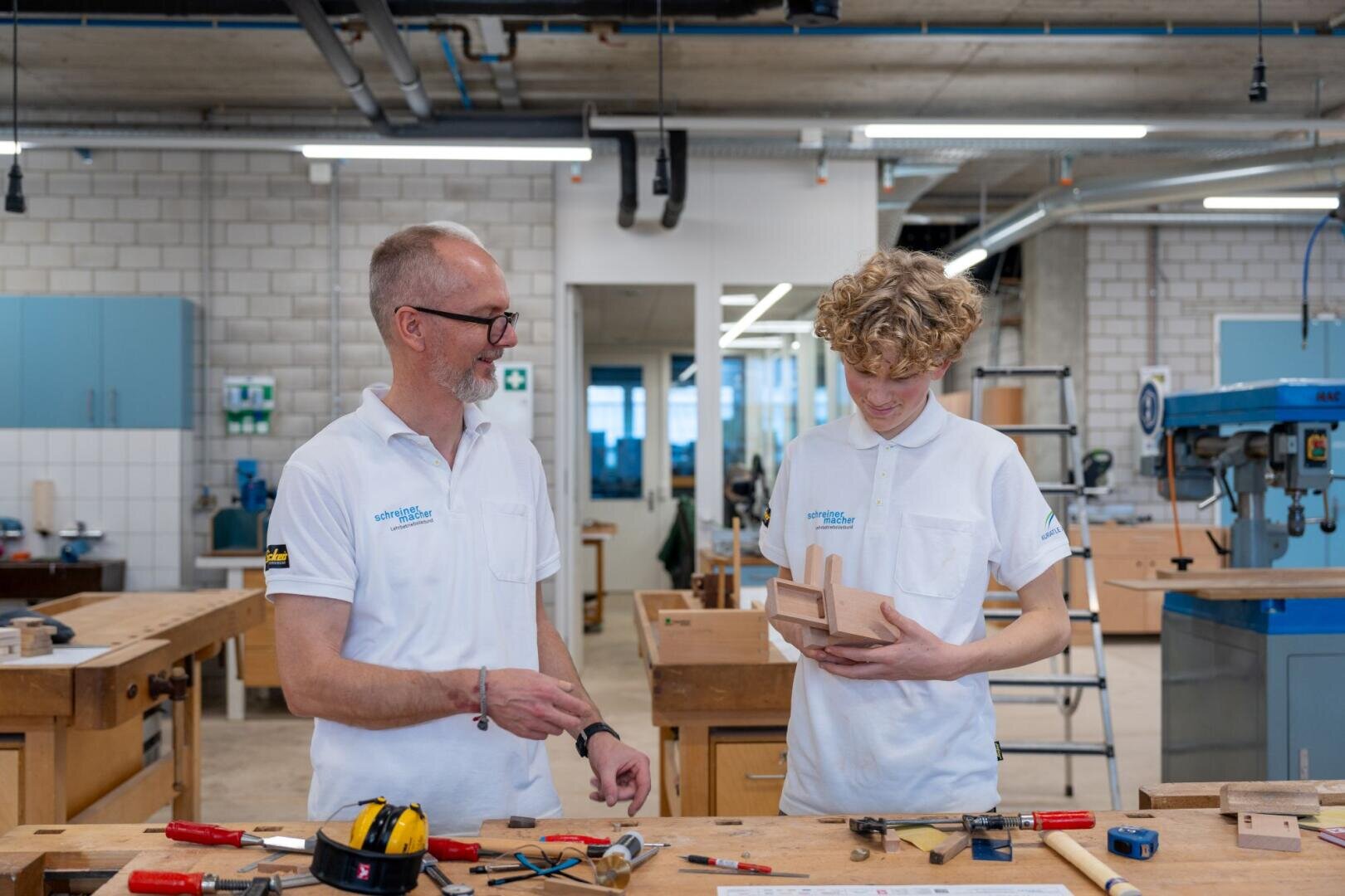 Zwei Personen in weißen Polohemden stehen in einer Werkstatt. Der eine, ein älterer Mann, lächelt, während er sich mit einer jüngeren Person unterhält, die ein Holzprojekt begutachtet. Im Hintergrund sind Werkbänke, Werkzeuge und Holzbearbeitungsgeräte zu sehen.