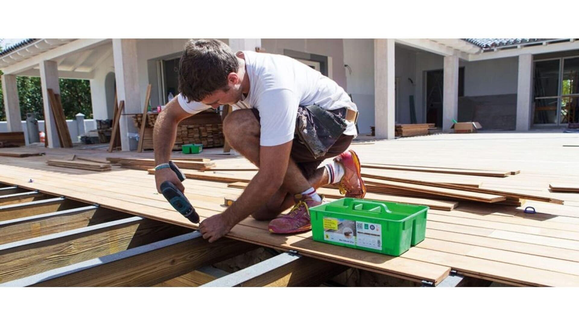 Un homme en short et tee-shirt blanc utilise une perceuse électrique pour installer une terrasse en bois à l'extérieur d'une maison. Des planches de bois et une boîte à outils verte se trouvent à proximité, la terrasse partiellement achevée étant visible.