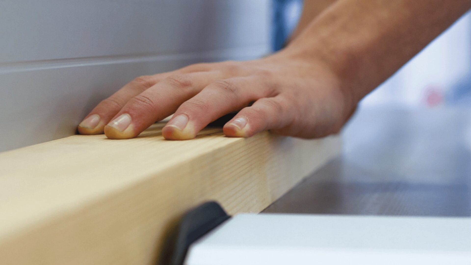 Nahaufnahme einer Person, die ein Holzstück auf einer Tischsäge oder Holzbearbeitungsmaschine führt, wobei der Schwerpunkt auf sorgfältiger Handhabung und handwerklichem Können liegt.