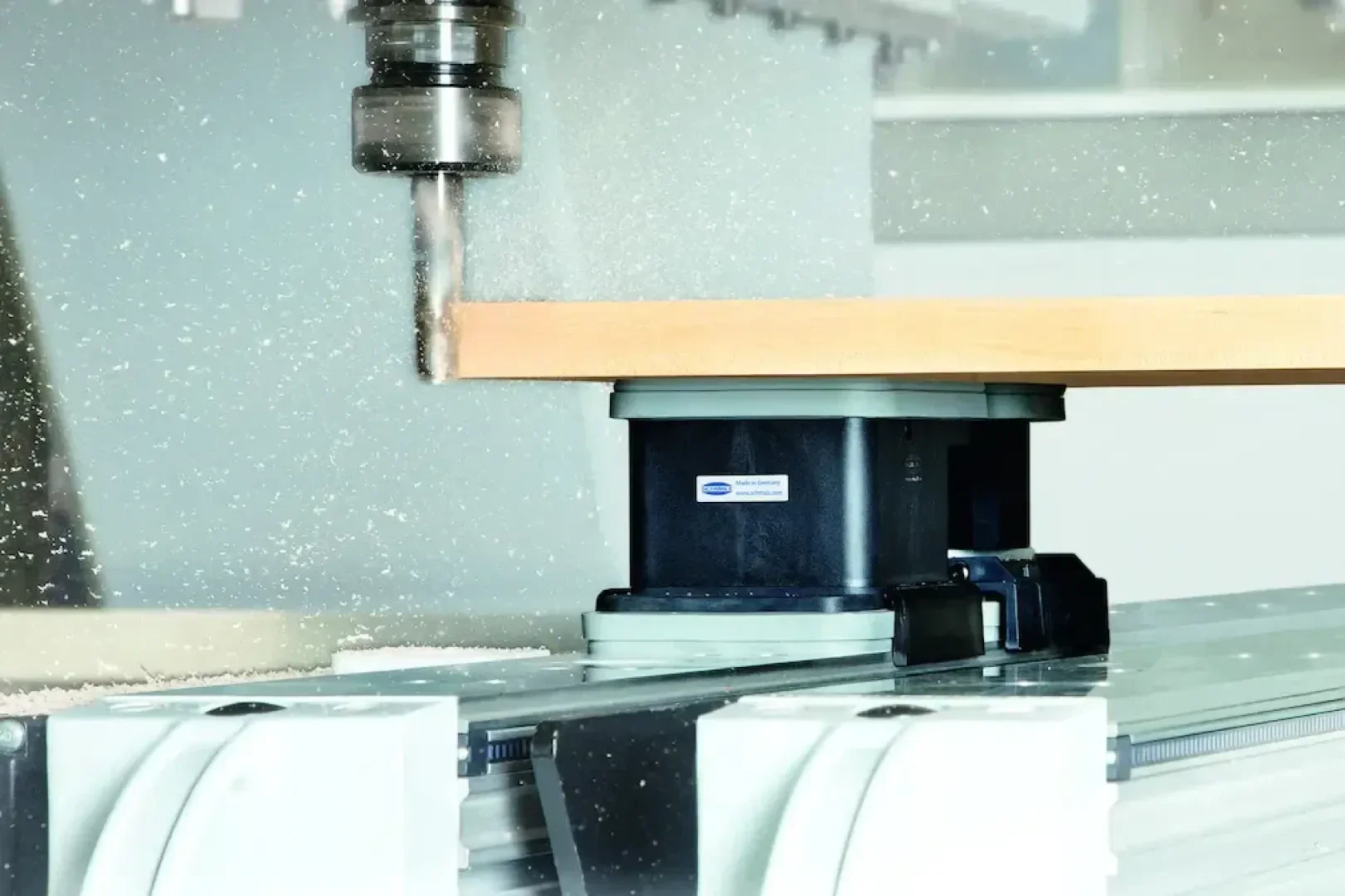 Eine CNC-Maschine bohrt in ein Holzbrett, wobei Sägespäne entstehen. Das Brett wird mit einem Saugnapf auf einem Metall-Arbeitstisch gehalten.