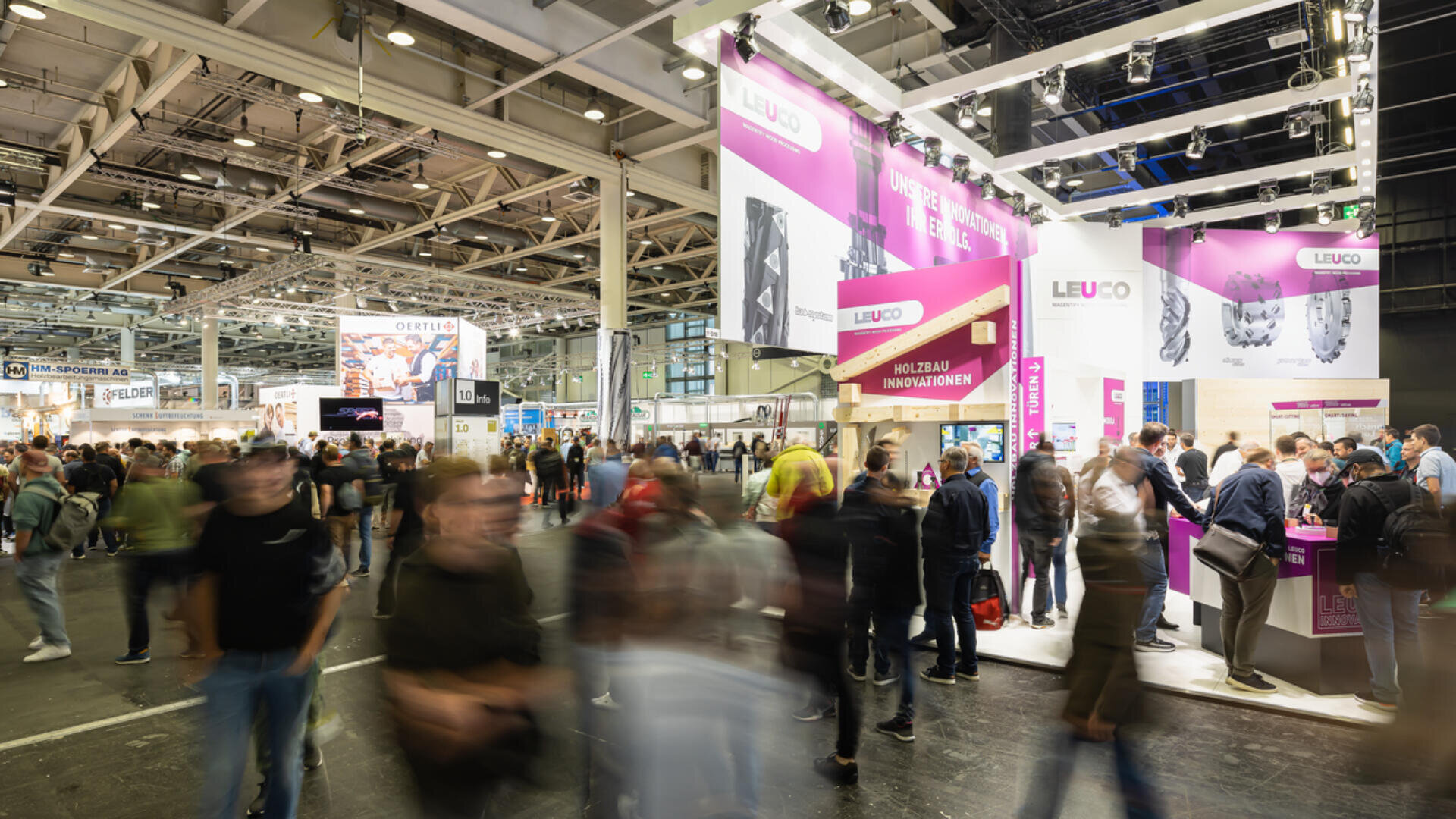 Ein belebtes Messegelände mit vielen Menschen, die die hell erleuchteten Messestände besuchen; über einem zentralen Stand sind auffällige rosa und weiße LEUCO-Schilder zu sehen.