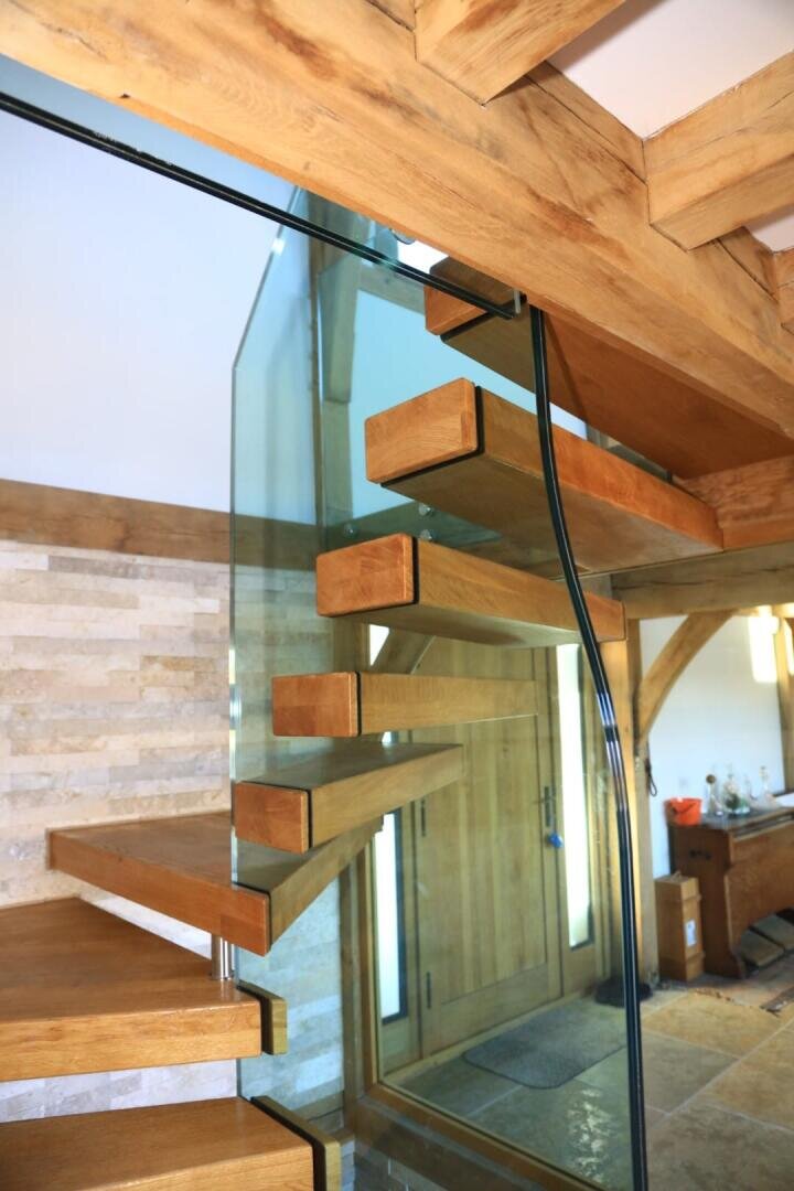 Eine moderne Holztreppe mit schwimmenden Stufen und Glasgeländer in einem Raum mit Holzbalken, Steinwänden und einer Holztür im Hintergrund.