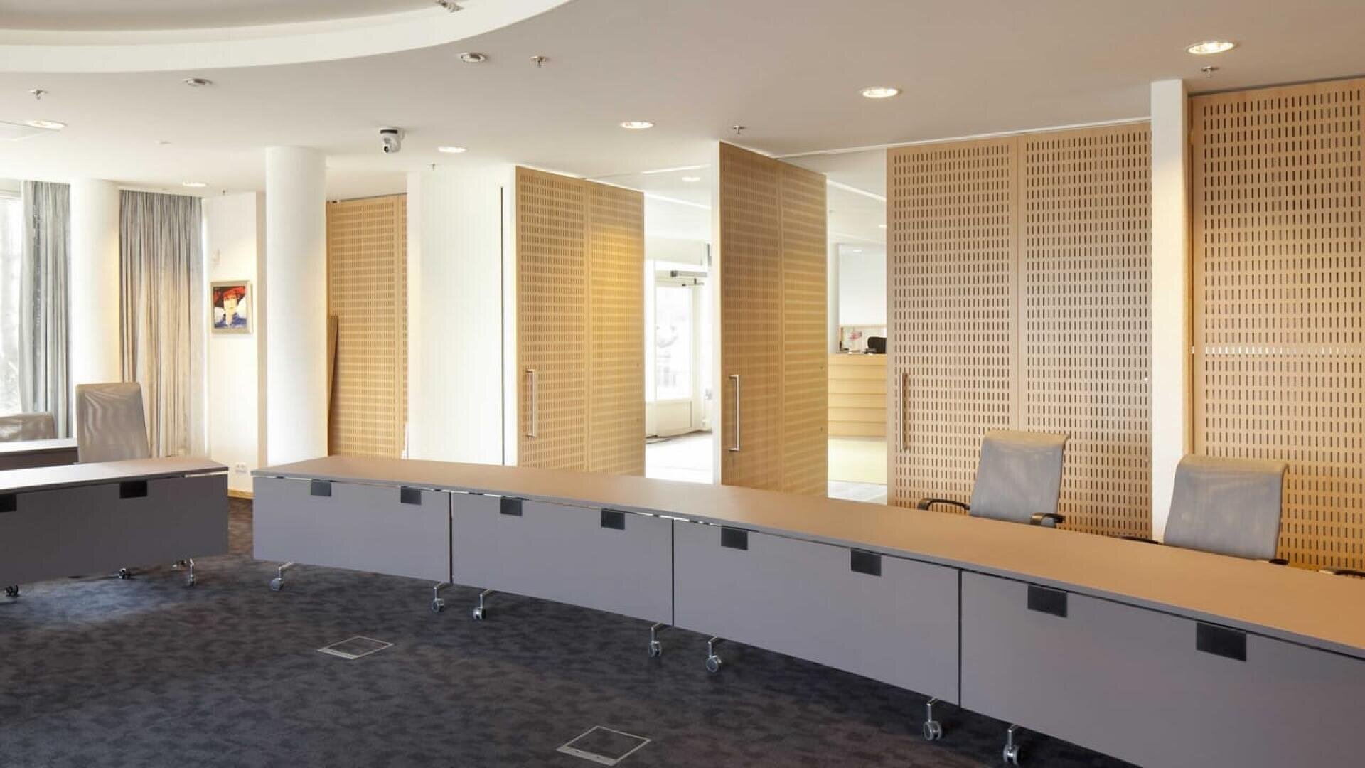 Une salle de conférence moderne avec une table incurvée sur roulettes, des chaises de bureau grises, des panneaux en bois clair et de grandes fenêtres laissant entrer la lumière naturelle. Des portes de séparation sont partiellement ouvertes à l'arrière-plan.