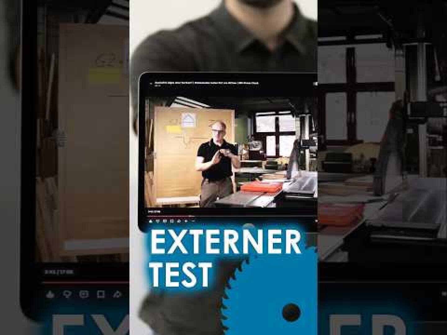 Eine Person hält ein Tablet in der Hand, auf dem ein Video läuft, das jemanden in einer Werkstatt zeigt. Auf dem Bildschirm ist „EXTERNER TEST“ zu sehen, darunter ein blaues Zahnrad-Symbol. Im Hintergrund sind Werkzeuge und Werkstattgeräte zu erkennen.