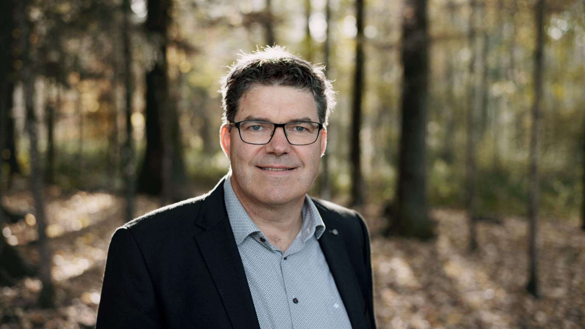 Ein Mann, der eine Brille, ein graues Hemd und einen dunklen Blazer trägt, steht im Freien in einem sonnenbeschienenen Wald und lächelt in die Kamera. Der Hintergrund ist unscharf, man sieht hohe Bäume und heruntergefallene Blätter.