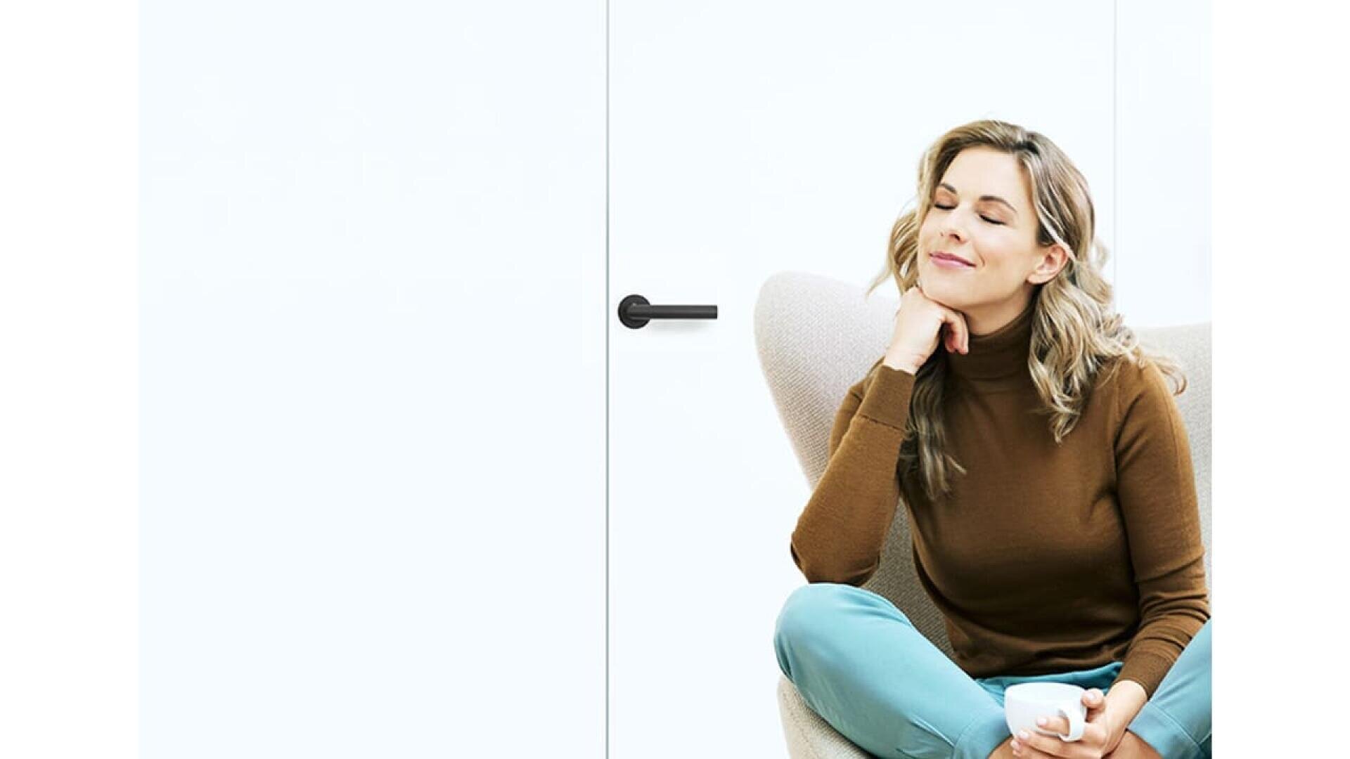 Une femme aux cheveux blonds ondulés, portant un col roulé marron et un pantalon sarcelle, est assise dans un fauteuil moderne, les yeux fermés et le sourire satisfait, tenant une tasse blanche à la main, sur un fond blanc.