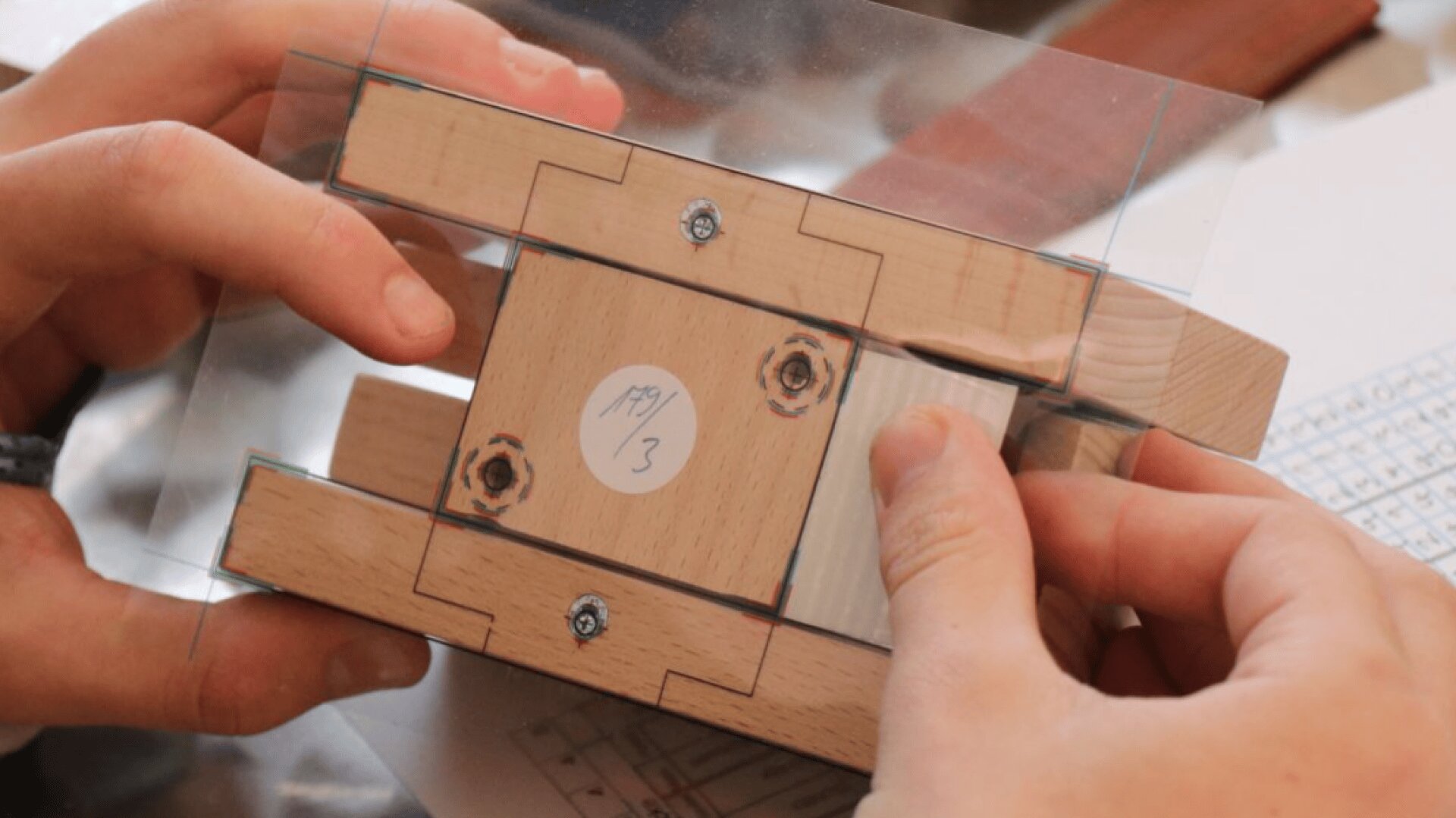 Les mains tiennent une petite structure en bois et en acrylique avec des vis et un autocollant étiqueté 139/3, peut-être une partie d'un modèle ou d'un prototype mécanique, à côté d'une feuille de papier avec une grille imprimée.