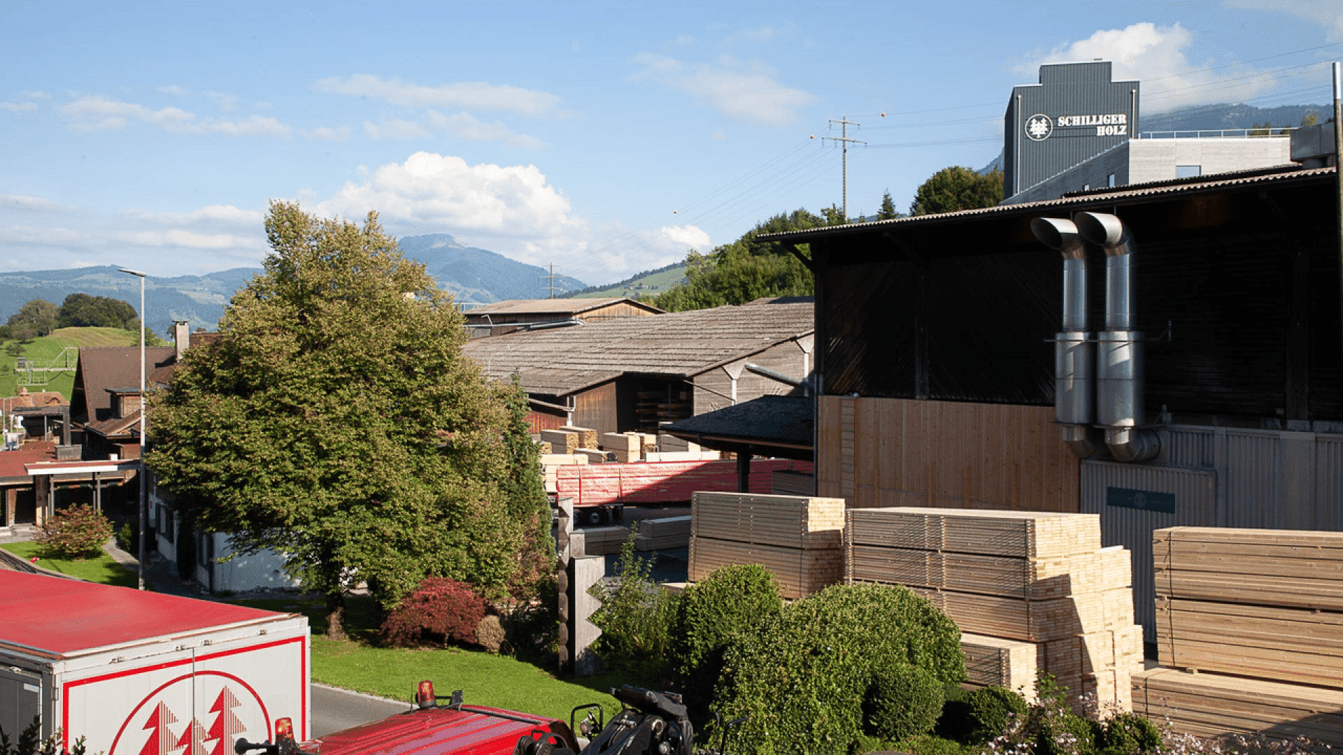 Un chantier forestier avec des piles de planches de bois, des bâtiments industriels et un camion au premier plan. Des arbres et des montagnes sont visibles à l'arrière-plan sous un ciel partiellement nuageux.