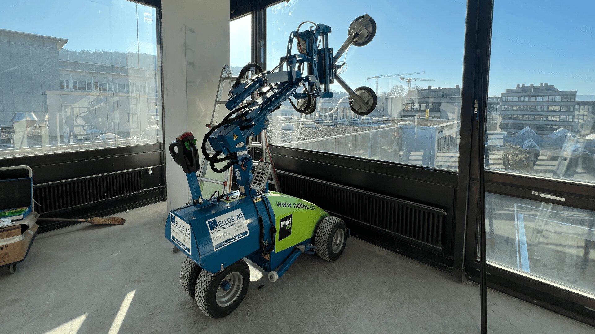 Ein blau-grüner Fensterinstallationsroboter mit Saugnäpfen steht im Inneren eines Gebäudes in der Nähe großer Fenster, wobei Baugeräte und Gebäude von außen zu sehen sind.