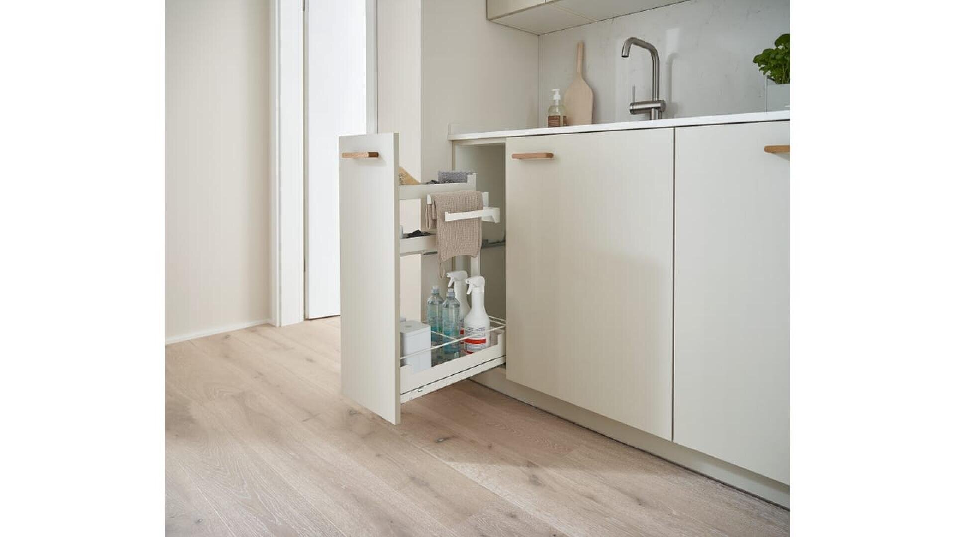 Une cuisine moderne avec des planchers en bois clair et des armoires blanches comporte un tiroir coulissant sous l'évier, rangé avec des produits de nettoyage, des chiffons et des flacons pulvérisateurs. Une plante verte est posée sur le comptoir.