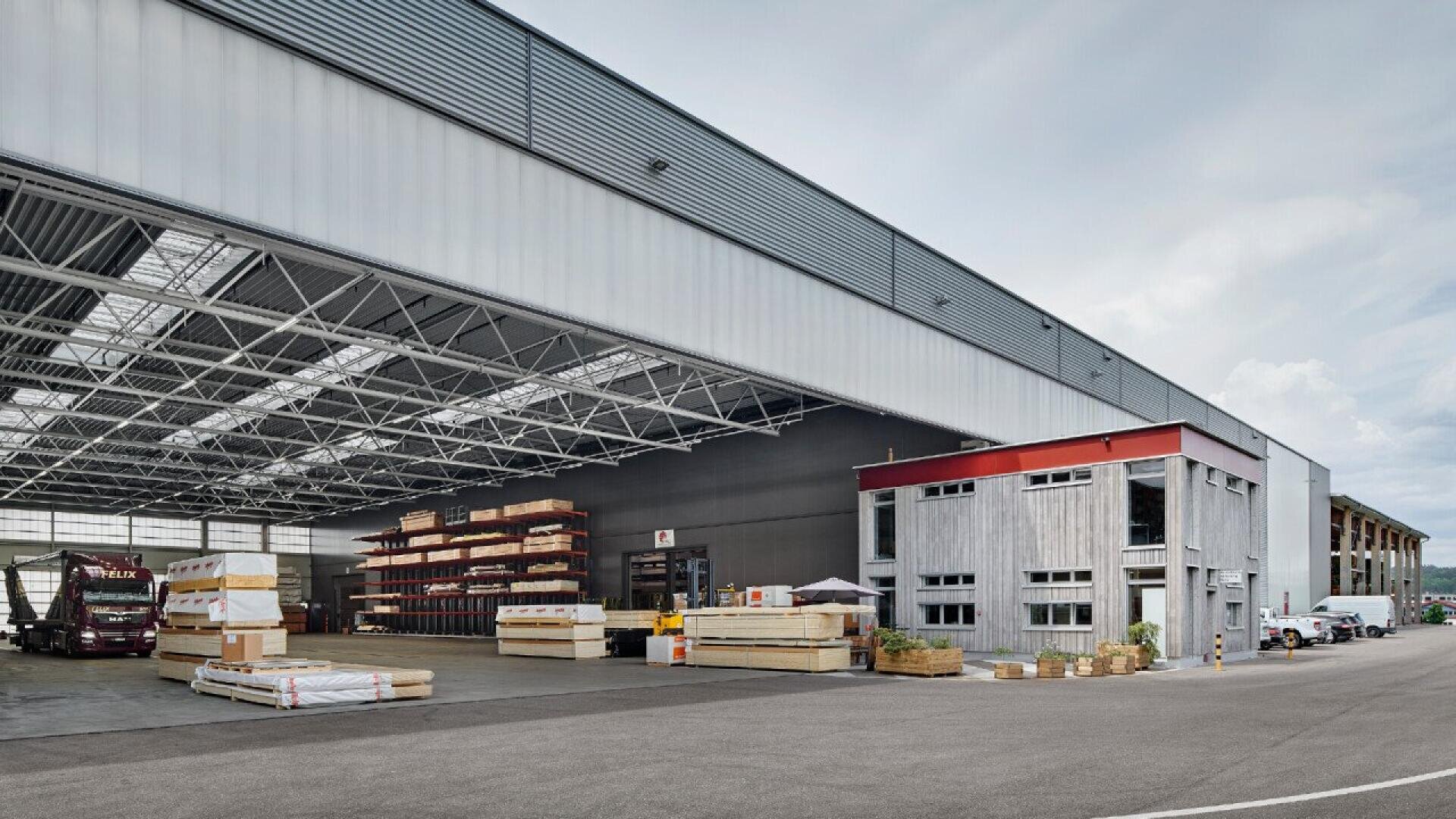 Un grand entrepôt industriel avec une zone de chargement ouverte, du bois empilé, une section de bureaux sur deux étages, des camions de transport garés à proximité et un ciel dégagé au-dessus de la tête.