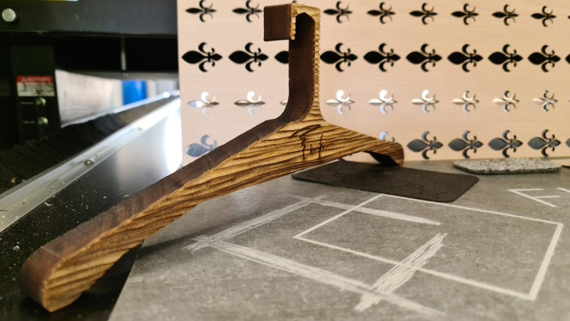 Ein hölzerner Kleiderbügel mit einzigartigem, eckigem Design steht auf einer grauen Fläche. Im Hintergrund befindet sich eine dekorative Tafel mit einem sich wiederholenden Fleur-de-Lis-Muster und verschiedenen Materialien.