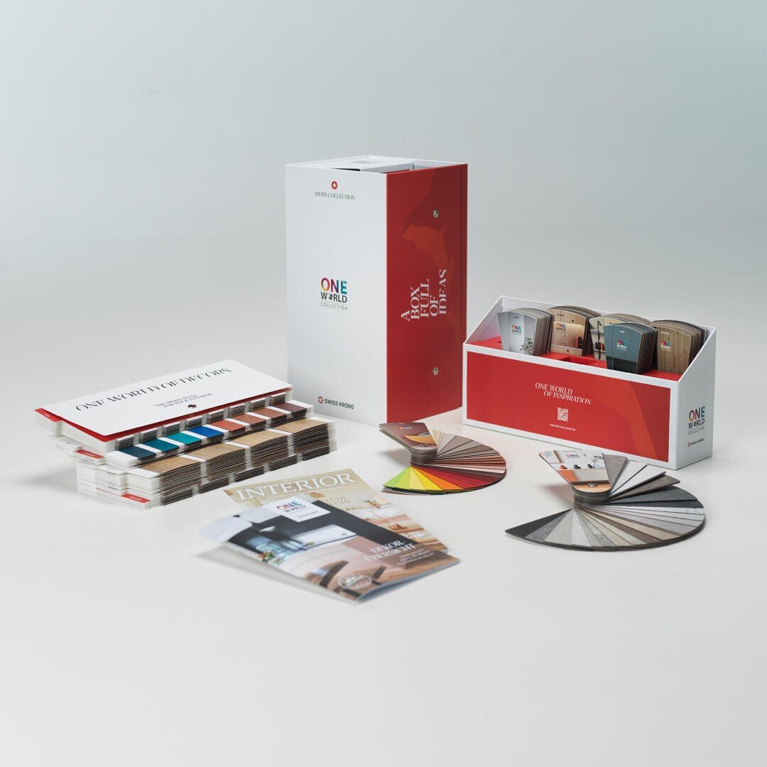 Ein Display mit Bodenbelagsmusterkits, Farbmustern, einer aufgeschlagenen Broschüre und einer Zeitschrift, die auf einer weißen Fläche mit weißem Hintergrund angeordnet sind. Die Artikel zeigen verschiedene Farben und Muster für die Innenraumgestaltung.