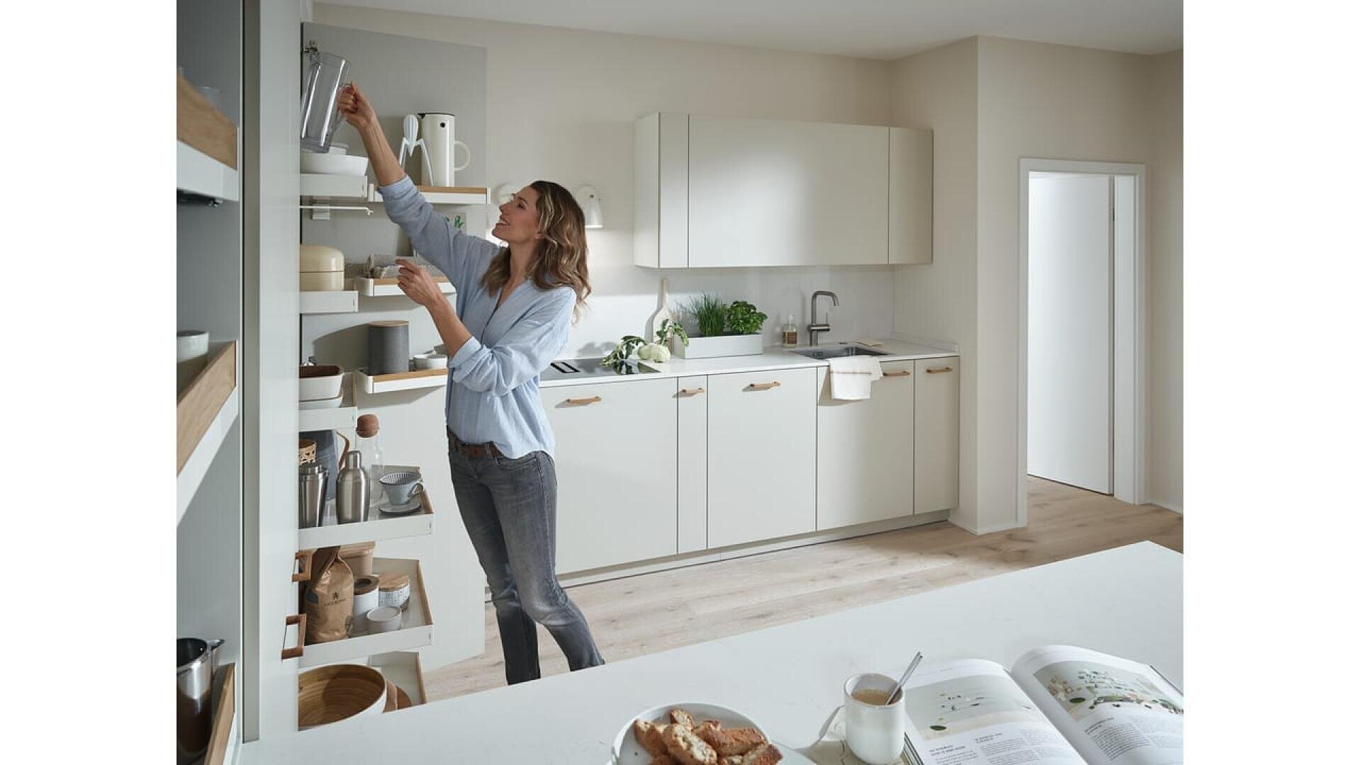 Une femme se tient dans une cuisine moderne et lumineuse, cherchant un objet sur une étagère en hauteur. Un livre de cuisine ouvert, du café et des pâtisseries sont posés sur le comptoir au premier plan. La cuisine présente des armoires blanches et des touches de bois.