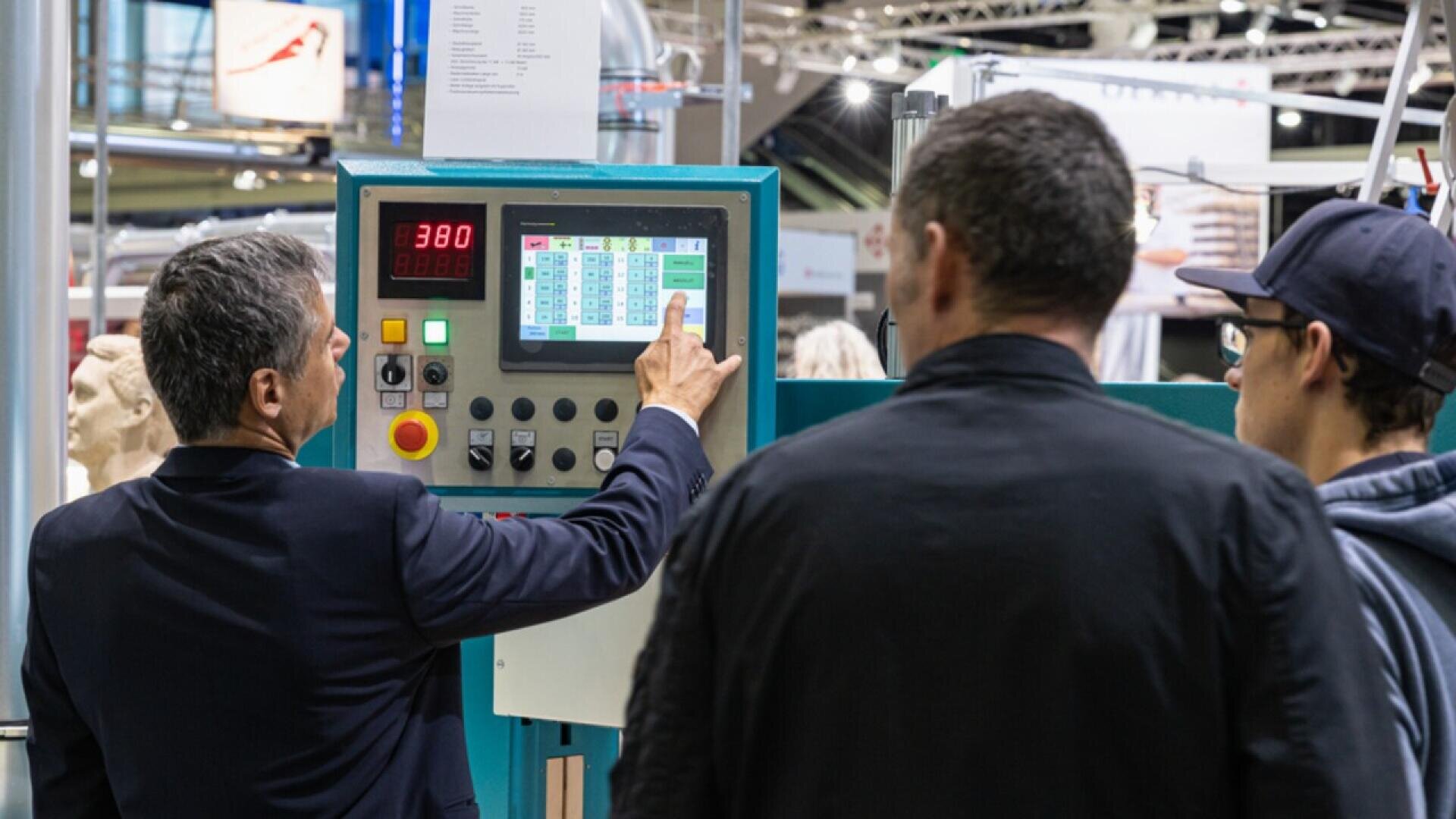 Trois hommes se tiennent devant un panneau de contrôle industriel doté d'écrans numériques et de boutons. Un homme en costume pointe l'écran tactile, expliquant quelque chose aux deux autres hommes qui font face à l'écran.