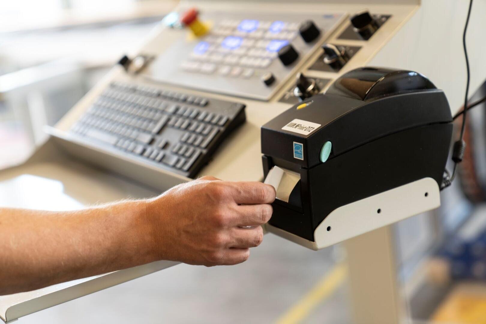 Eine Hand zieht ein Etikett aus einem schwarzen Thermo-Etikettendrucker an einem Industriearbeitsplatz. Im Hintergrund sind eine Tastatur und ein Bedienfeld mit Tasten und Reglern zu sehen.
