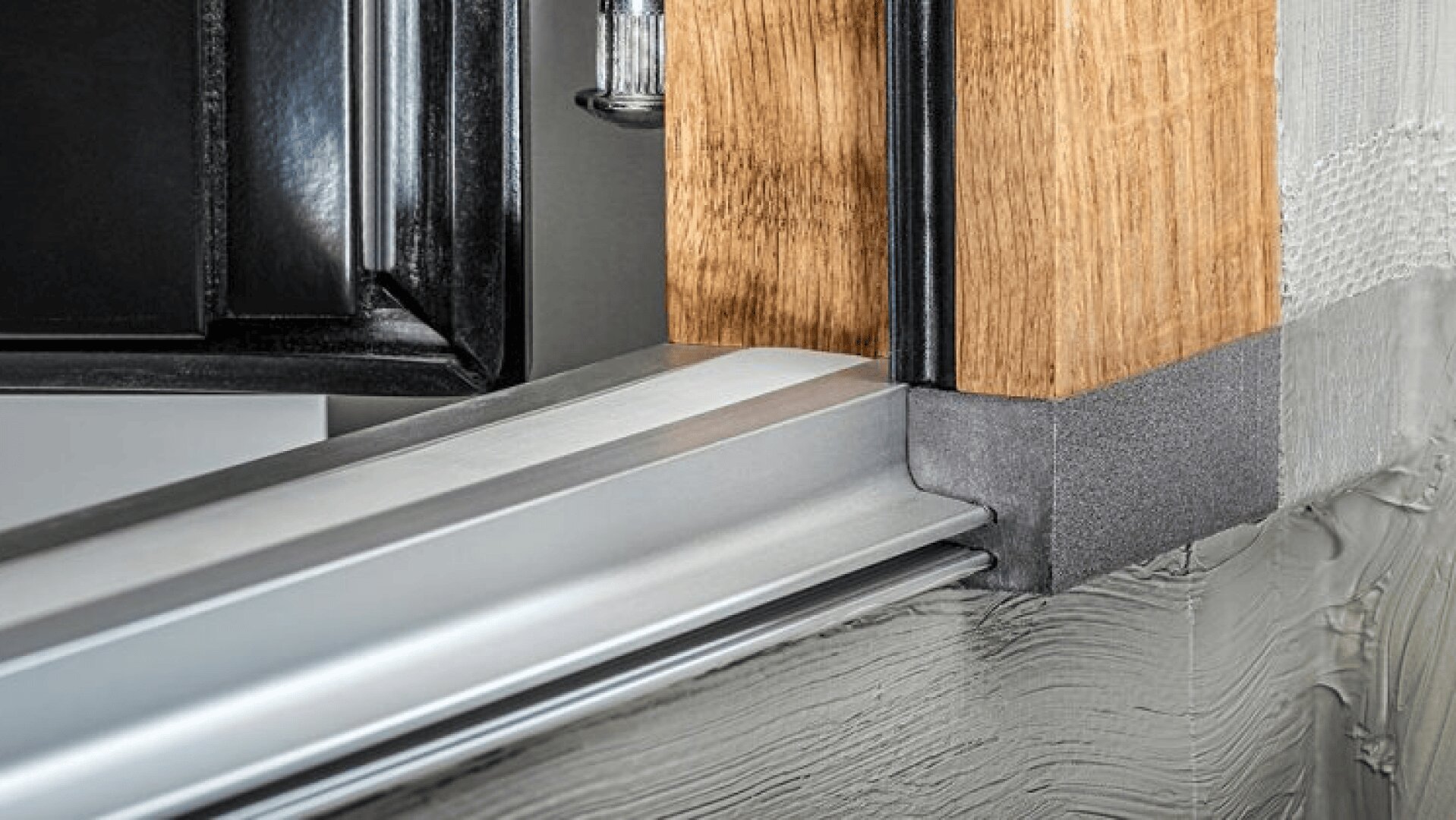 Nahaufnahme einer Fensterrahmenecke mit einem schwarzen Metallrahmen, einem Holzteil, einer silbernen Metallschwelle und einer grauen Dichtungsmasse, die zur Isolierung und Abdichtung auf die umgebende Fläche aufgetragen wurde.