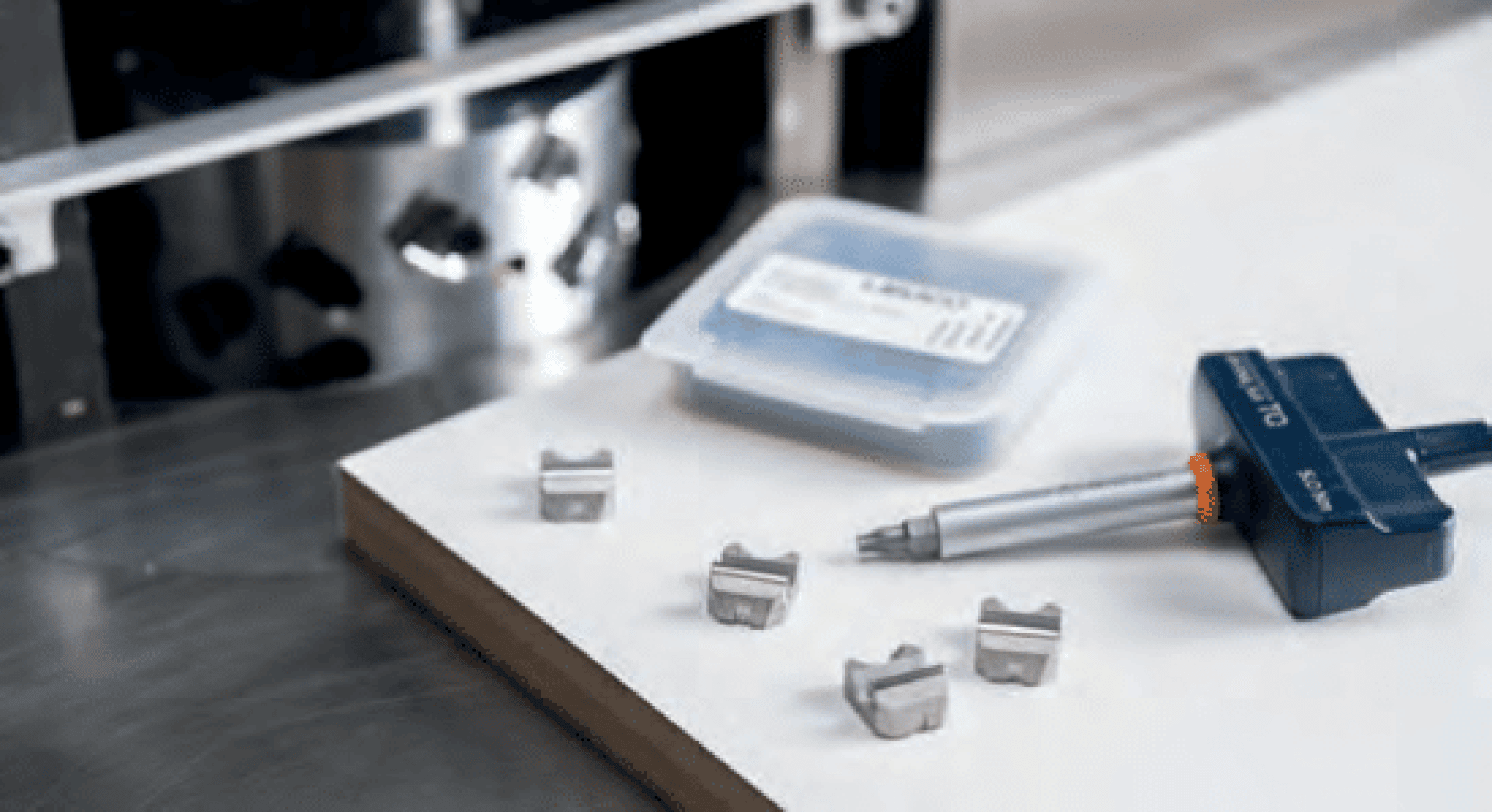 Ein digitaler Messschieber, drei kleine Maschinenteile aus Metall und ein Kunststoffbehälter stehen auf einer weißen Fläche in einer Werkstatt oder einem Labor.