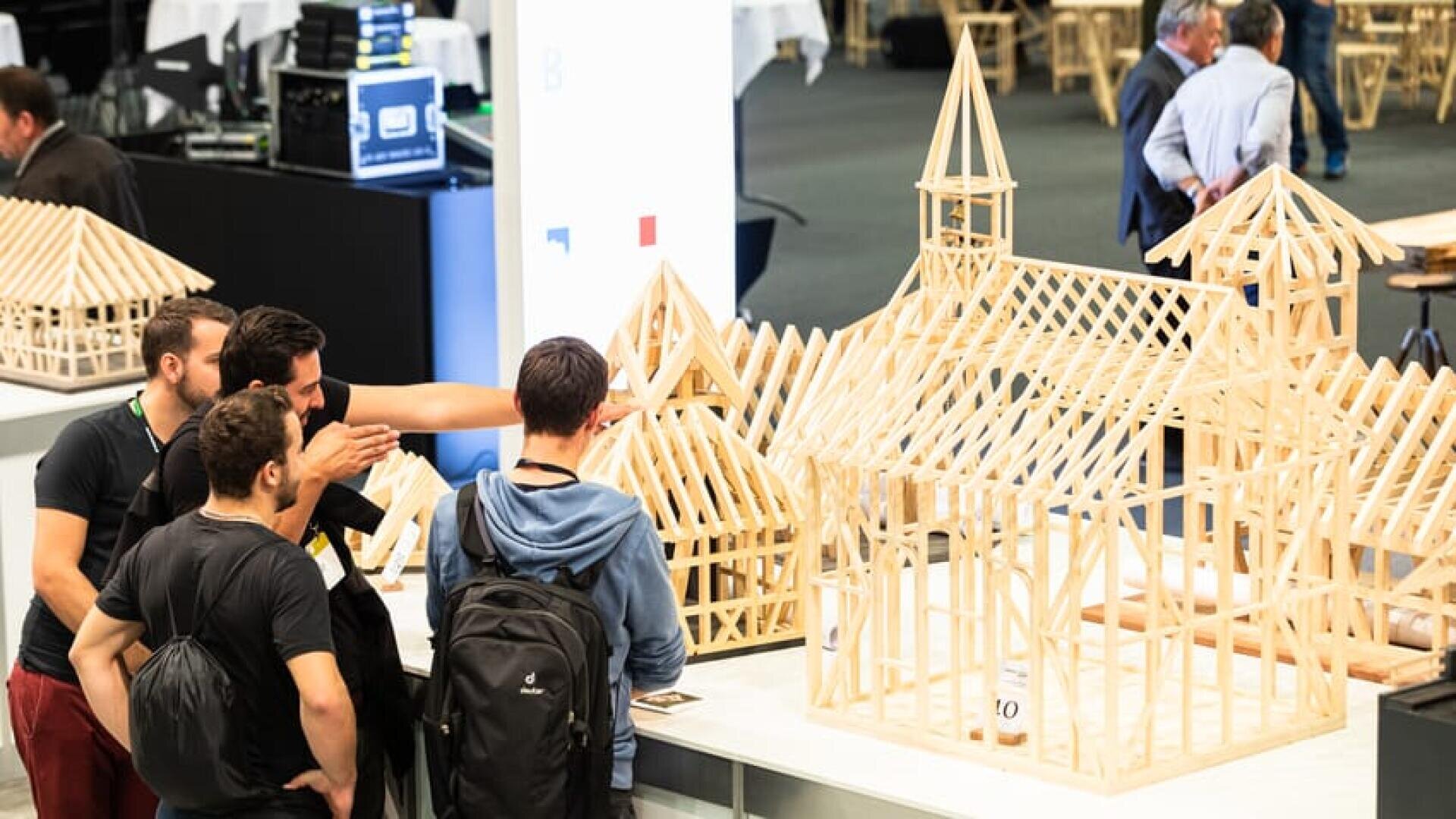 Eine Gruppe von vier Personen versammelt sich um einen Tisch und begutachtet auf einer Indoor-Veranstaltung oder Ausstellung detaillierte hölzerne Architekturmodelle von Dachstühlen und Gebäuden.