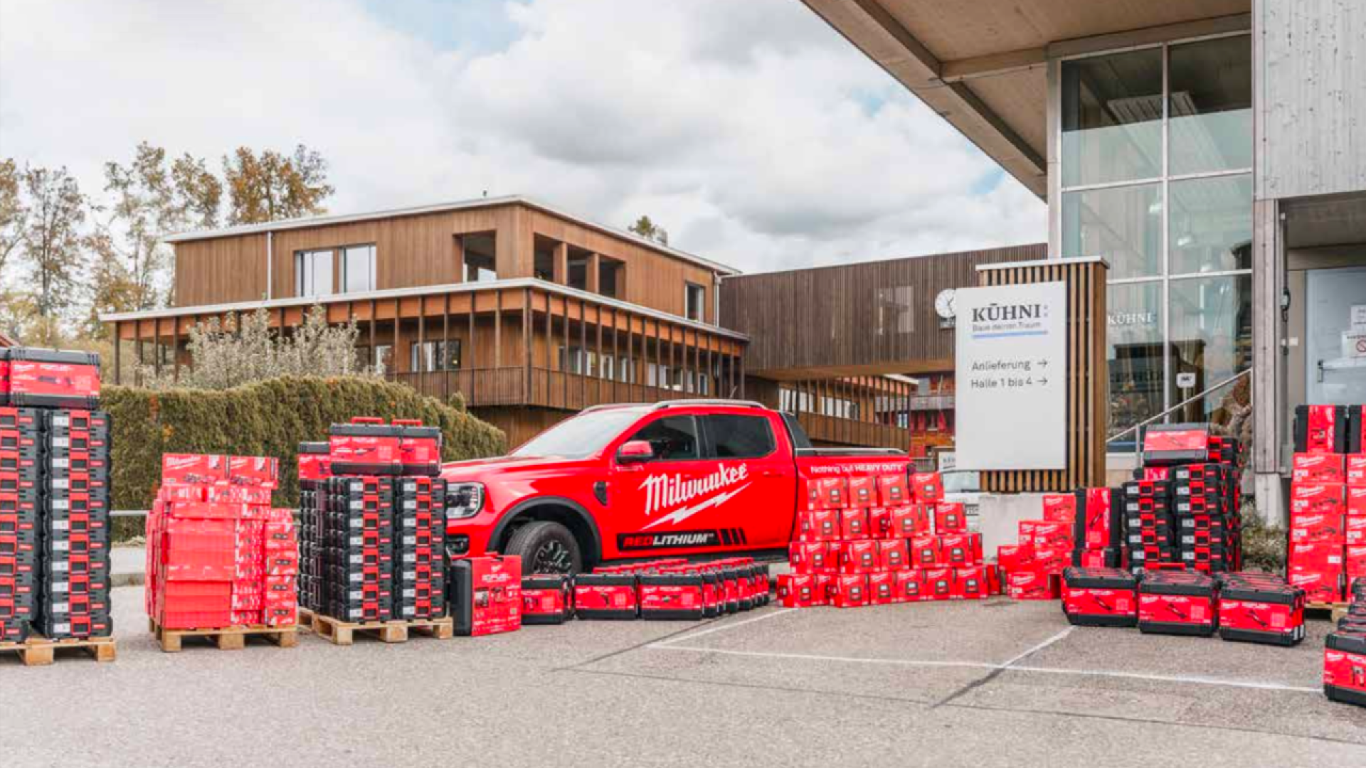 Une camionnette rouge de la marque Milwaukee est garée à l'extérieur d'un bâtiment moderne, entourée de nombreuses piles et palettes de boîtes à outils et d'équipements Milwaukee rouges et noirs. Des arbres et un ciel nuageux se trouvent à l'arrière-plan.