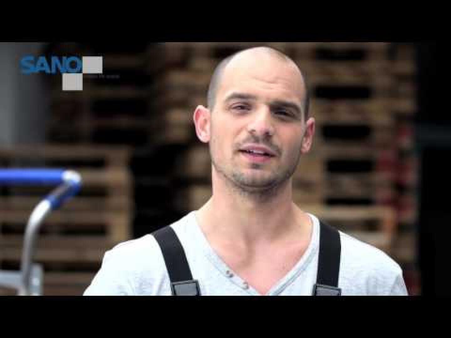 Un homme au crâne rasé et à la barbe courte, portant une chemise gris clair et des bretelles noires, se tient devant une pile de palettes en bois. Dans le coin supérieur gauche, on peut voir le mot SANO.