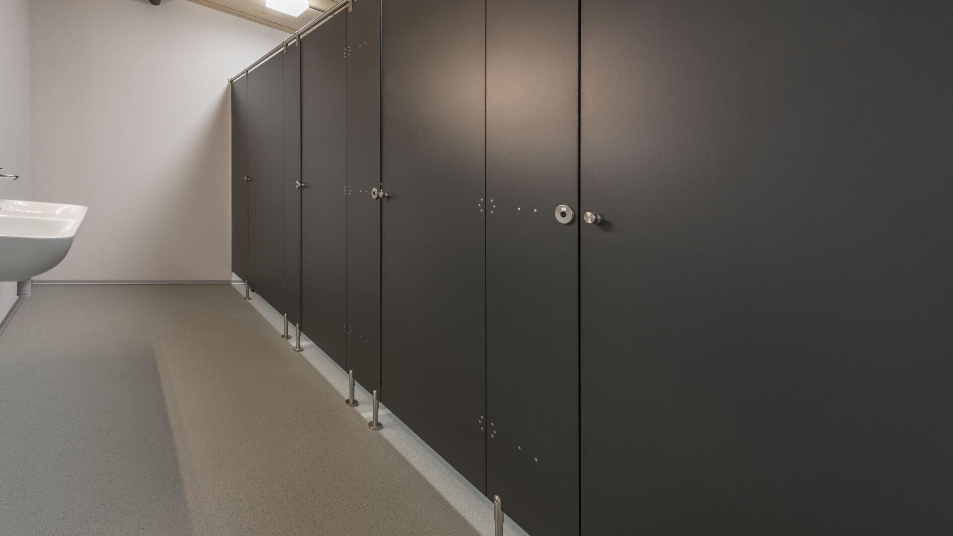 Eine Reihe geschlossener grauer Toilettenkabinen mit Metallschlössern steht in einer sauberen öffentlichen Toilette. Links an der Wand ist ein weißes Waschbecken angebracht, und der Boden ist mit hellen, gesprenkelten Fliesen ausgelegt.